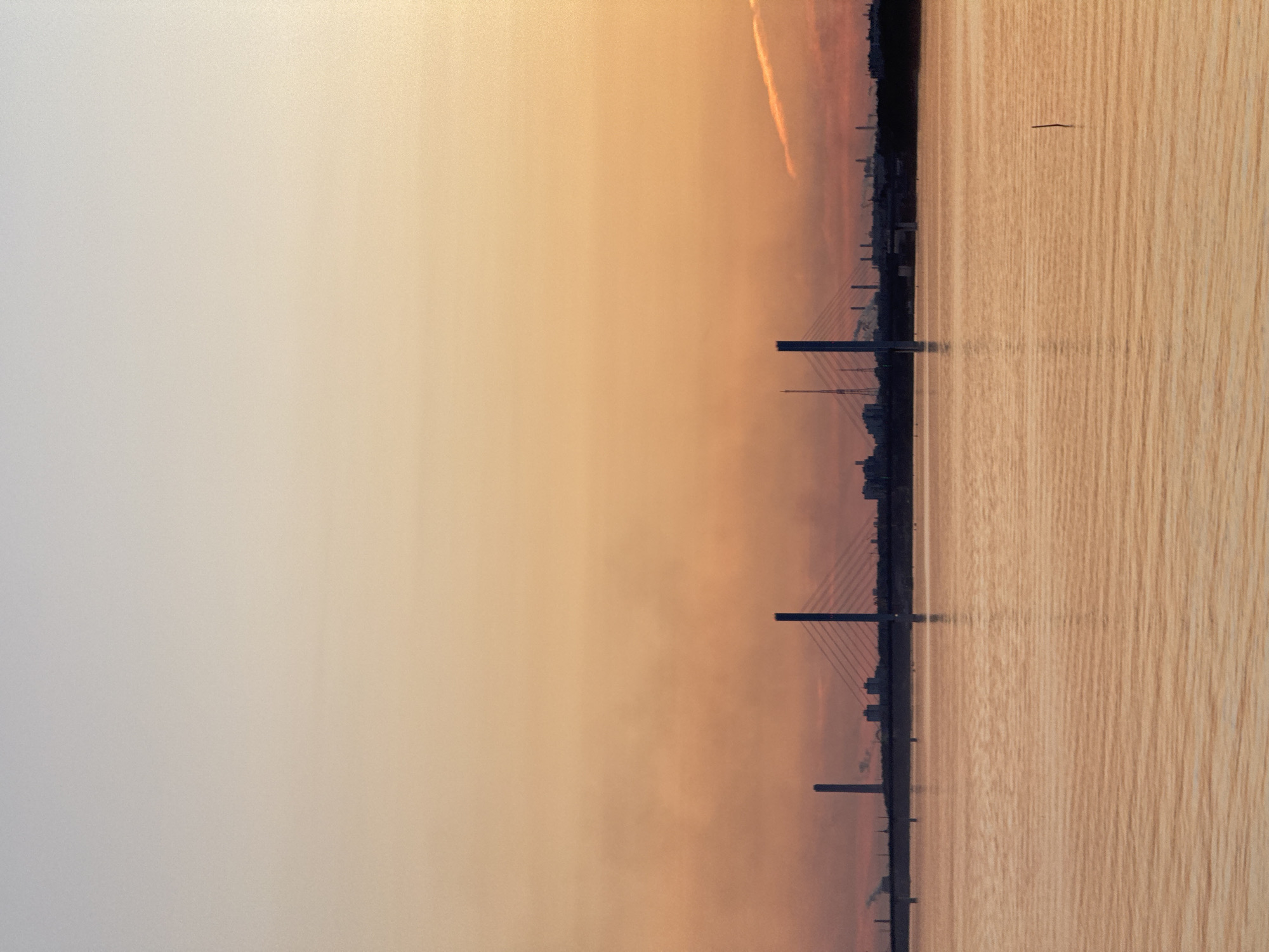 The image depicts a wide body of water, likely a river or bay, reflecting the warm hues of a sunset. The sky transitions from a pale, almost white at the top to a soft orange and pink gradient towards the horizon. Silhouetted against the colorful sky is a city skyline featuring industrial structures, smokestacks, and prominent bridges. One particularly large cable-stayed bridge stands out with its tall pylons and intricate network of cables. There are no people visible in the image. The water's surface is slightly rippled, creating a textured sheen that echoes the colors of the sky. A thin, dark vertical object, possibly a marker or a lone post, is present in the foreground of the water. The overall scene suggests an industrial port city at dusk.