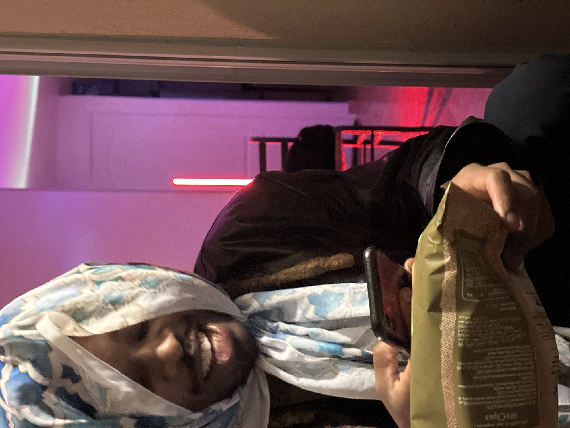 The image depicts a person wrapped in a blue and white patterned scarf, obscuring much of their face except for their mouth and teeth, indicating a smile. The person is holding a smartphone in one hand and a bag of chips in the other. A second person, partially visible, is wearing a dark hooded garment. The setting appears to be indoors, with pink and red lighting in the background. Through a window, a blurred scene suggests an exterior view. While I can discern the elements in the picture, I cannot determine its specific location in Montreuil, France, as it lacks landmarks or other indicators that would enable location identification.