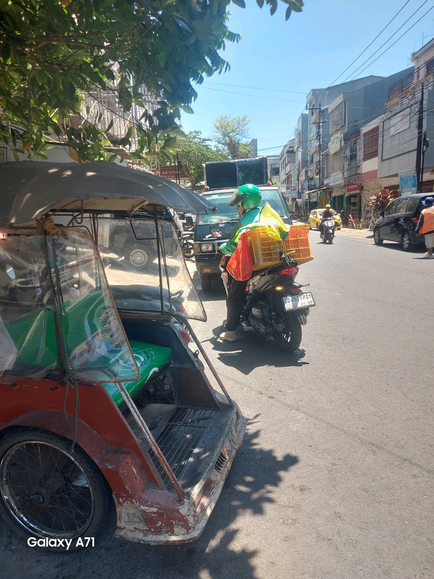 In Makassar, Indonesia, a street scene unfolds under a bright, clear sky. A three-wheeled motorcycle, commonly known as a becak, is parked on the left, its weathered red and silver body partially obscured by foliage. The becak has clear plastic sheeting covering the passenger area, revealing green seating inside. On the right, a person wearing a bright green helmet and a turquoise jacket is riding a motorcycle. They are carrying a yellow plastic crate filled with items, possibly for delivery, secured to the back of their motorcycle. Behind them, traffic flows along the asphalt road, with buildings lining the right side of the street. The buildings appear to be commercial, with signs and awnings visible. The overall atmosphere is that of a busy urban environment on a sunny day.