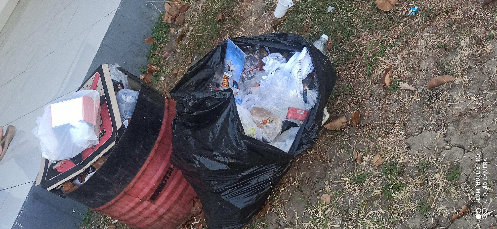 A black trash bag filled with garbage is on the ground next to a red and black metal barrel. The trash bag is bulging and appears to be overflowing. The barrel is also filled with trash, including a white plastic bag containing a pink and white rectangular object, possibly a box. Leaves and dry grass are scattered around the barrel and the trash bag on the ground, suggesting an outdoor environment. The ground appears to be a mixture of soil and patchy grass. A white disposable cup is visible to the right of the trash bag, and a plastic water bottle is near the bottom of the trash bag. The scene is set outdoors, with a tiled surface visible in the upper left corner, potentially indicating a walkway or patio area adjacent to the grassy area. The image was captured in Betro, Indonesia.