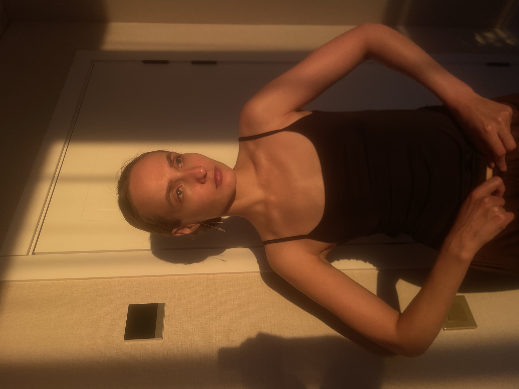 The image depicts a woman in a dark, sleeveless top, standing against a textured wall with a doorway and a light fixture. Golden light, characteristic of sunset or sunrise, casts strong shadows on the wall and the woman. Her hair is pulled back, and she is looking to her right. Her hands are positioned near her waist, as if adjusting her clothing. A shadow of the woman is visible on the wall behind her, elongated by the low angle of the light. The environment appears to be an indoor setting, possibly a hallway or a room, with the light source originating from outside the frame. The location in the city of Подшивалово, Russia, is not discernible from the visual information provided in the image.