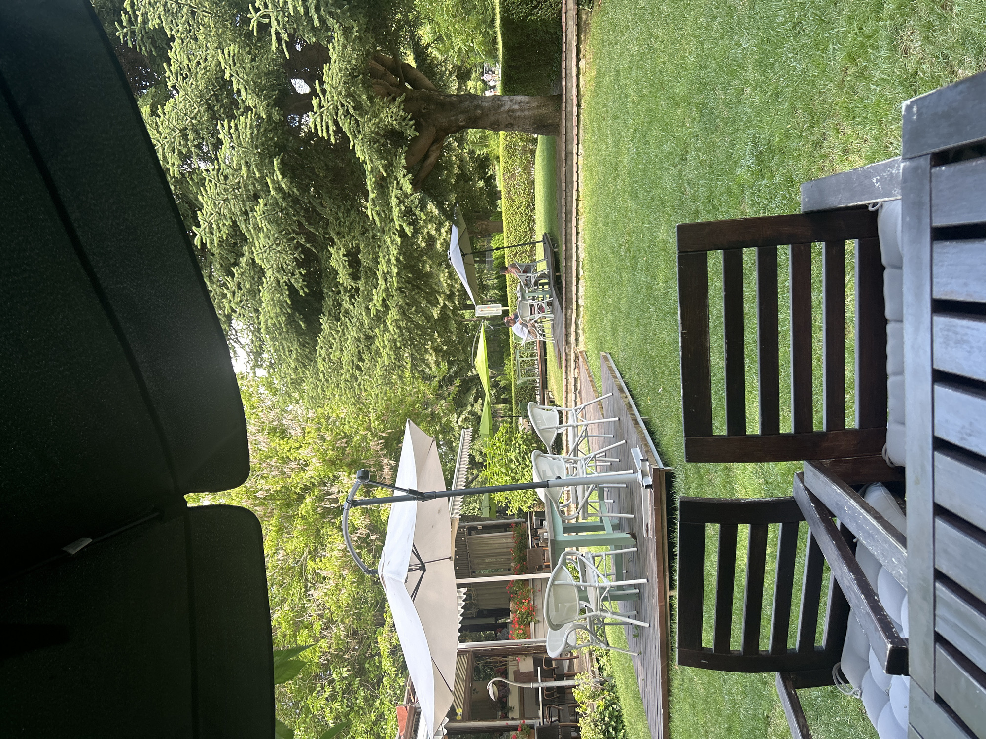 The image depicts an outdoor seating area of a restaurant or cafe, likely in Varna, Bulgaria, given the presence of abundant greenery and a large mature tree that suggests a well-established park or garden setting. In the foreground, two dark wooden chairs with light grey cushions are positioned on a wooden deck, facing away from the camera and towards a lush green lawn. A portion of a dark wooden table is also visible in the extreme foreground.
Beyond the foreground chairs, a wooden deck area hosts a light blue table surrounded by four white wicker chairs, all situated beneath a large white umbrella with a curved grey pole. This area appears to be set up for dining. In the background, a white building with awnings and large windows is visible, with more outdoor seating and umbrellas in front of it. Further into the distance, people are seated at tables under smaller green umbrellas, and a well-maintained hedge forms a backdrop. A very large, mature evergreen tree dominates the right side of the frame, its branches extending over the seating area. The environment is verdant and appears to be a sunny day.