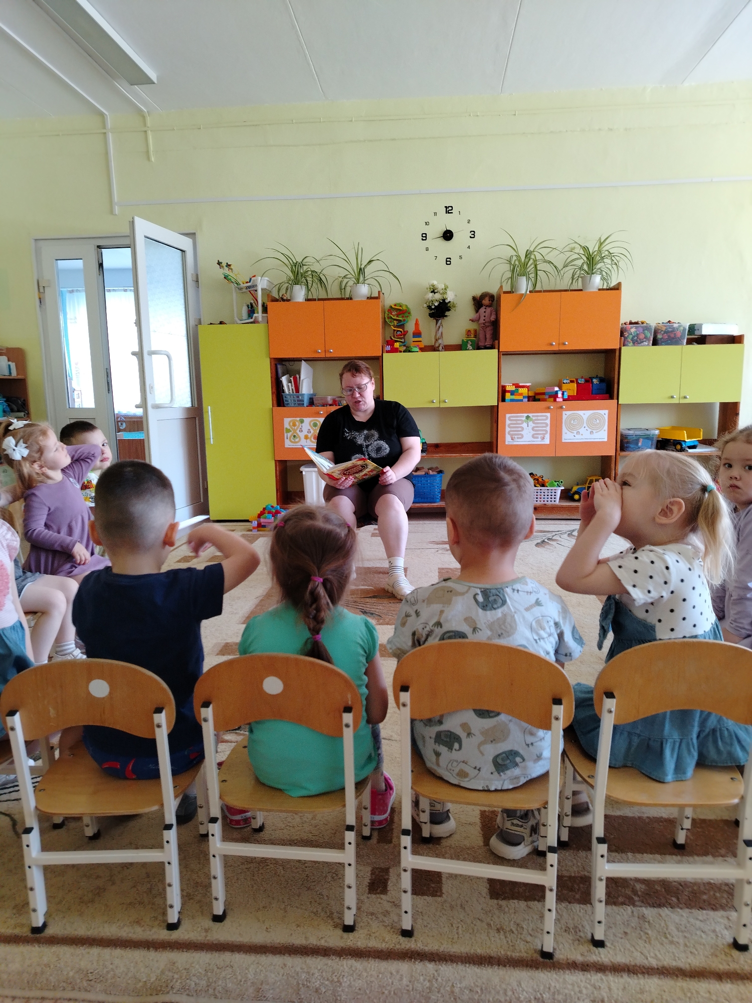 A woman is reading a book to a group of young children. The children are sitting on small wooden chairs, arranged in a semi-circle facing the woman. The woman is seated on a larger chair. Behind her is a brightly colored cabinet with toys, plants, and decorations. A clock is mounted on the wall. The location is a classroom or play area in what is described as the city of Chaikovsky, Russia.