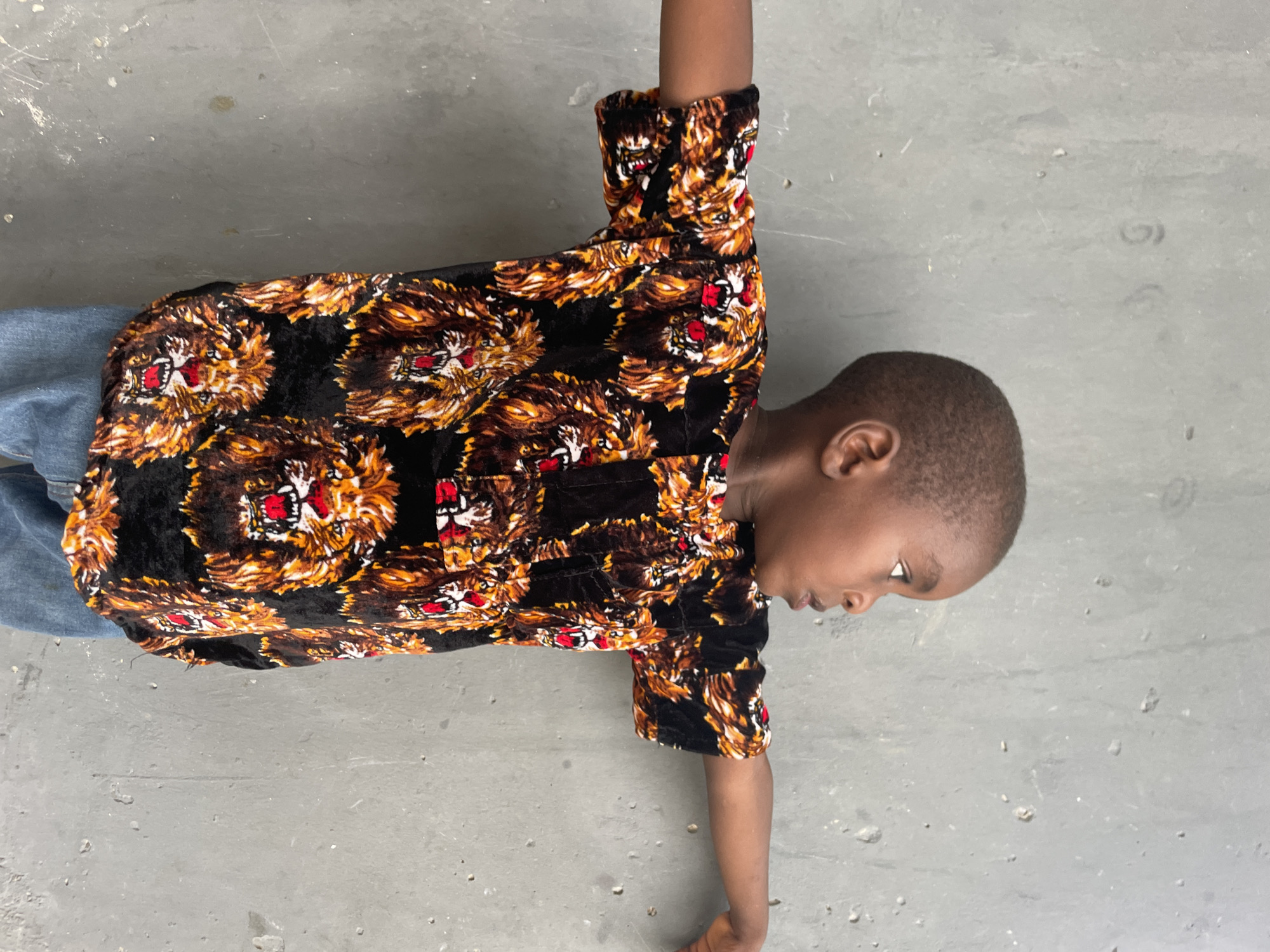 A young person, likely a child, is seen lying or posing on a light grey, possibly concrete, surface. They are dressed in a long-sleeved shirt featuring a distinctive, repetitive pattern of animal faces, resembling lions or tigers, with prominent orange and brown fur details accented by red and white around the eyes and mouths, all set against a black background. Blue denim jeans are visible on their lower body.
The individual has short dark hair and is positioned with their head turned to their right (viewer's left), looking downwards. Their right arm is extended upwards, and their left arm extends downwards, with the hand resting on the ground. The pose appears casual or playful. The grey surface beneath is somewhat rough and shows minor scuffs and marks. The lighting is bright and even, indicating daytime. There is no visible text in the image. The setting suggests a simple, functional space in Badagry, Nigeria.