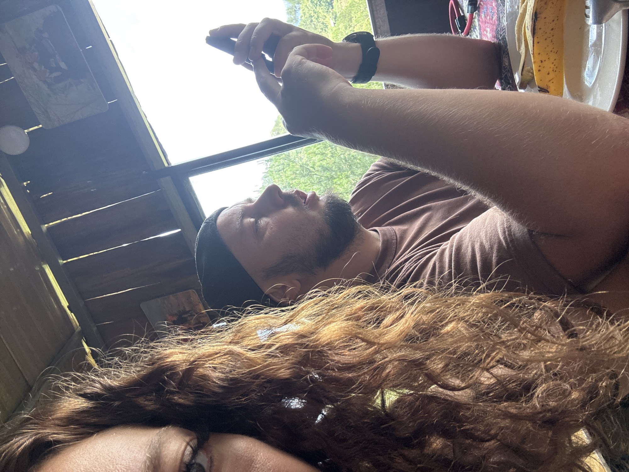 A close-up, tilted shot shows two people indoors, likely in Ngã Ba Kim, Vietnam, based on the setting's style. One person, a male, is in the background wearing a brown t-shirt, black hat, and has a beard. He is holding a smartphone. A watch is visible on his wrist. The second person, mainly visible due to their brown, curly hair, is in the foreground with only the left eye visible. The setting has wooden planks, and a window looks out onto trees. A plate with a banana peel and cutlery is also present.