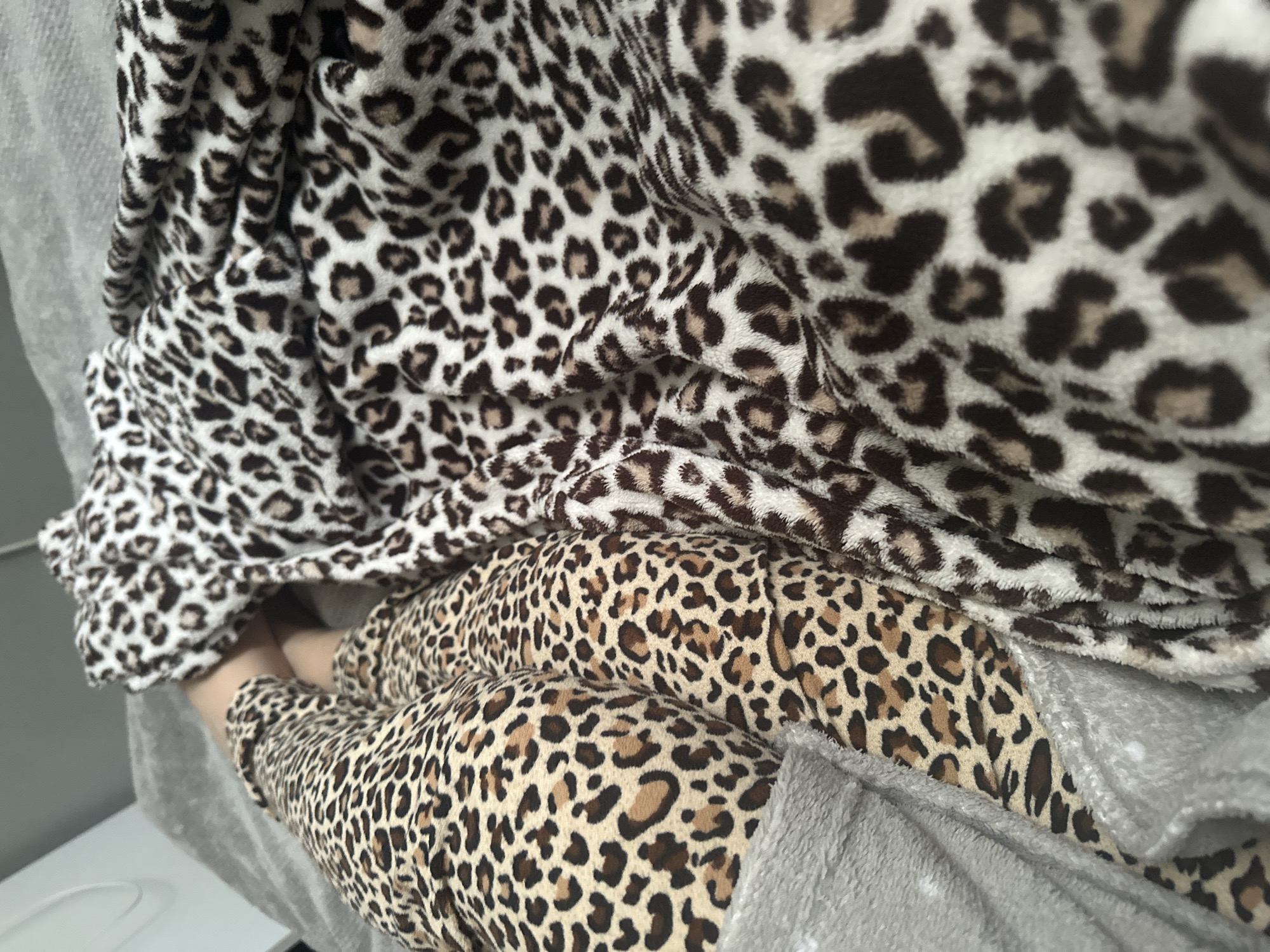 The image captures a close-up, high-angle perspective of a person's lower body resting in a reclined position. The individual's legs are encased in a pair of light brown and dark brown leopard-print pants, fabricated from a soft, plush material. Their bare feet, displaying a light skin tone, are visible at the center, protruding from beneath the fabric. A distinct cream and dark brown leopard-print blanket, composed of a similar soft, plush texture, is draped over a portion of the legs and extends across the right side of the frame. Both leopard prints feature dark brown rosettes, with the pants exhibiting a tan base and the blanket a white/cream base with light brown accents within some rosettes. The subject and fabrics are supported by a textured, light grey fuzzy surface, likely a blanket or bedding. A blurred, light-toned vertical element is partially discernible in the upper left background, indicating an indoor setting in Unknown, Unknown.
