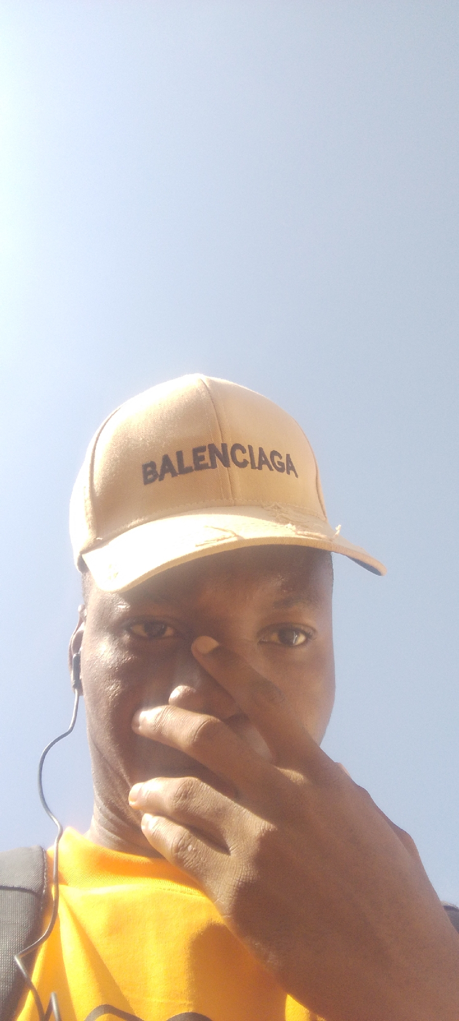 The media file shows a close-up, low-angle, outdoor selfie of a person wearing a beige baseball cap with "BALENCIAGA" printed in black letters. The person is also wearing a bright yellow t-shirt and headphones with black wires. Their right hand is raised, covering part of their face, with fingers obscuring their nose and mouth. The person's eyes are visible, and they appear to be looking towards the camera. The background is a clear, bright sky, suggesting it is daytime with good weather. The lighting indicates the sun is high in the sky. The setting appears to be outdoors, possibly in Nigeria given the location context. No specific activity or event is clearly depicted, but the selfie suggests the person is taking a photo of themselves. The overall mood appears casual and candid.