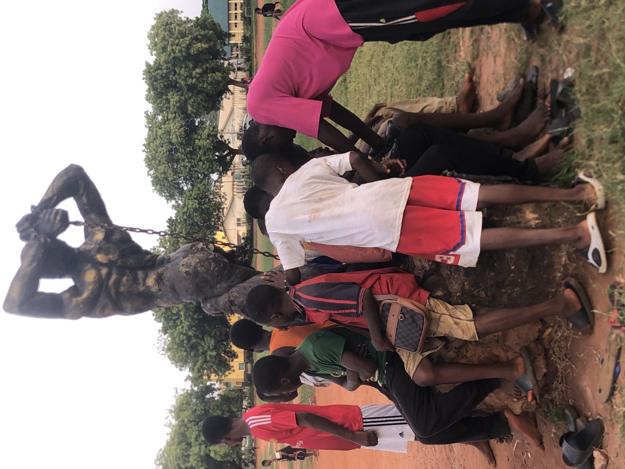 The image, taken in Nsugbe, Nigeria, captures a group of approximately ten to twelve young males, predominantly children and teenagers, gathered around a prominent, dark-colored statue. The statue depicts a muscular, partially clothed male figure with heavy chains attached to its wrists, suggesting themes of struggle or enslavement.
The scene is set outdoors in what appears to be a public area, possibly a park, school ground, or communal space. The ground consists of bare earth with some patches of grass. Several trees are visible in the background, along with distant buildings that blend into the landscape.
The boys are dressed in casual attire, including t-shirts, shorts, and track pants. One boy's shorts display the "ADIDAS" logo. They are congregated around the base of the statue, with some sitting or leaning on the ground, while others stand nearby. Their relaxed postures suggest they are resting, socializing, or observing their surroundings. One boy wears a small, patterned shoulder bag.
The lighting is soft and diffused, characteristic of a late afternoon, early evening, or an overcast day, casting no harsh shadows. There is no explicit activity taking place beyond the casual gathering of the youth around the significant statue. The overall impression is one of a quiet, reflective gathering in an outdoor community setting.