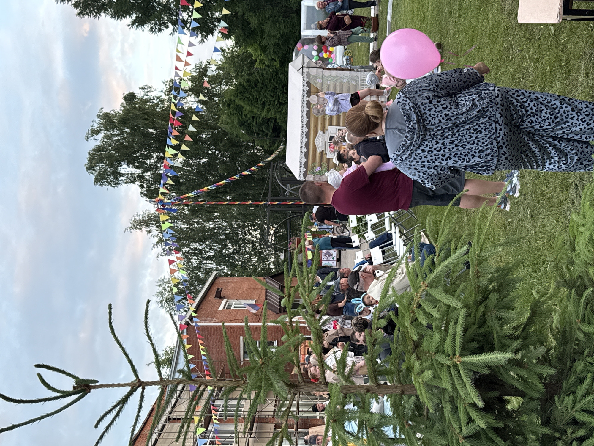 The image depicts an outdoor gathering in a village setting, possibly in the city of Kukuí, Russia, given the context provided. In the foreground, a large evergreen tree dominates the left side of the frame. Behind the tree, a crowd of people are assembled on a grassy area. Several individuals are seated on white folding chairs, facing a small, wooden structure decorated with what appears to be a painted facade resembling a traditional Russian izba.
A woman in a leopard-print tracksuit is walking away from the camera towards the right, holding a large pink balloon. A man in a burgundy t-shirt is holding a baby. In the background, a brick building with scaffolding is visible. Colorful triangular flags are strung across the scene, strung between poles and trees, suggesting a festive or celebratory atmosphere. The sky is overcast with some hints of color, indicating it might be late afternoon or early evening. Various trees and foliage are scattered throughout the background.