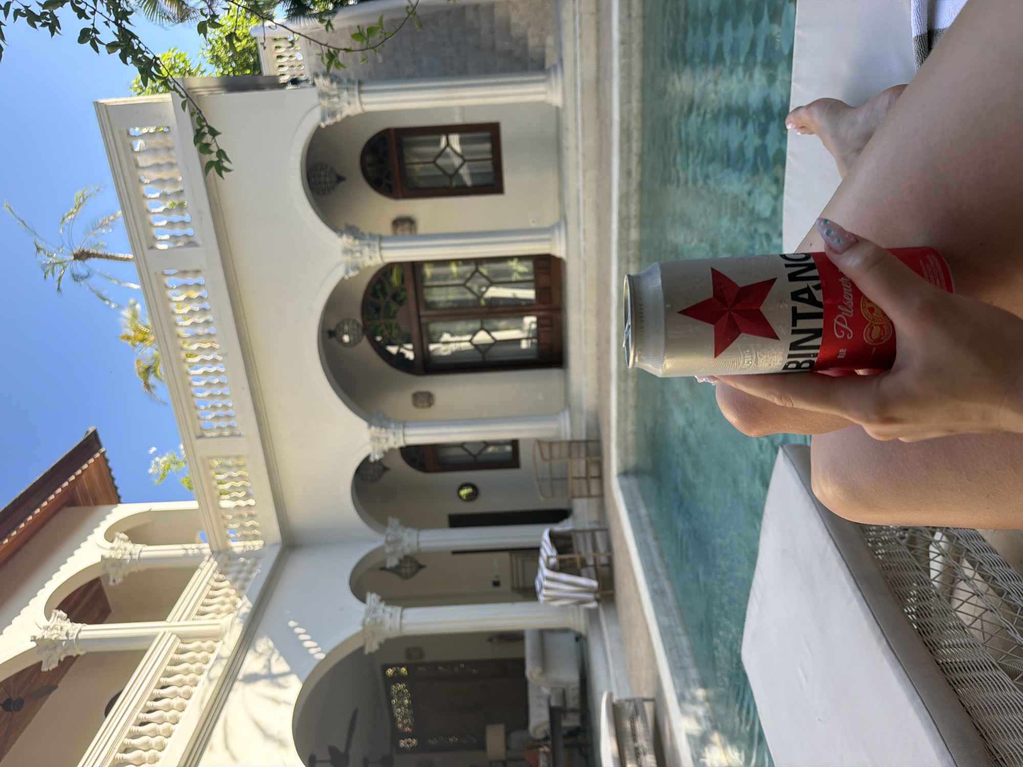 This image, presented from a first-person perspective and rotated 90 degrees clockwise, captures a moment of leisure by a pool in North Kuta, Indonesia. The scene is bathed in bright daylight under a clear blue sky, indicating warm and sunny weather.
The primary subject is a person, likely female, relaxing on a light-colored lounge chair beside a swimming pool. Only their legs, feet, and one hand are visible. The hand, with manicured nails featuring a subtle design, holds a silver can of Bintang Pilsener beer, identifiable by its prominent red star logo and "BINTANG Pilsener" text. Condensation on the can suggests it is cold.
The setting is a luxurious resort or villa, characterized by ornate white architecture with a Mediterranean or colonial influence. The building features multiple levels with arched openings, intricate balustrades, and decorative columns with classical capitals. Dark wooden doors and windows are visible within the arched recesses. In the background, palm trees and lush green foliage are seen against the vibrant blue sky, reinforcing the tropical location. A clear, light blue swimming pool runs along the side of the building, reflecting the sunlight. The overall impression is one of relaxation and vacation in a beautiful, upscale tropical environment.