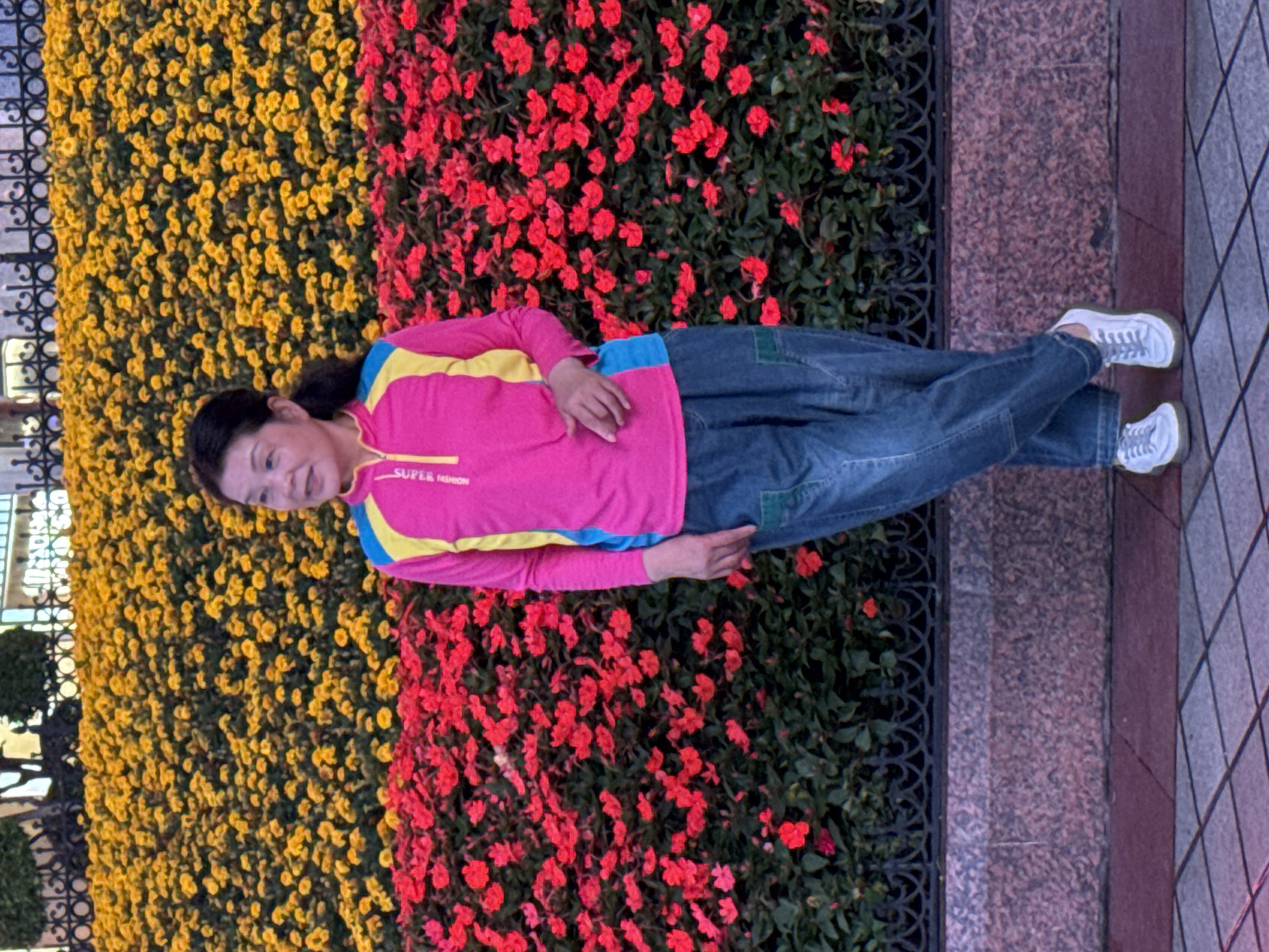 A woman is standing in front of a wall of flowers, with yellow flowers to her left and red flowers to her right. She is wearing a pink shirt with yellow and blue stripes, blue jeans with a green patch, and white sneakers. The location is likely Yuzhong District, China. The flowers are planted behind a black wrought iron fence that is affixed to a granite wall. The walkway is gray tile.