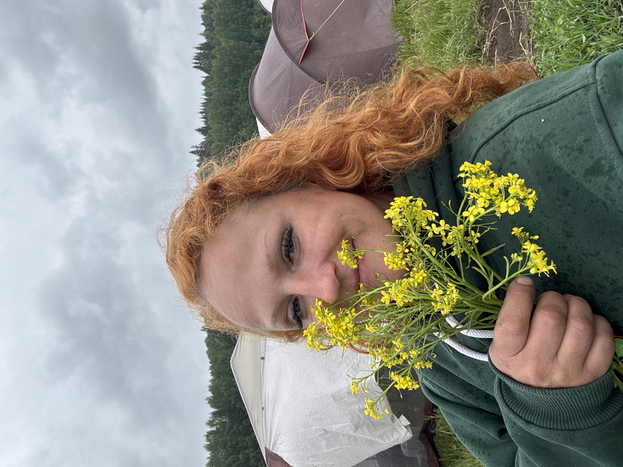 An outdoor portrait captured in Большая Кивара, Russia, shows a young woman holding a bouquet of small yellow wildflowers. The woman, with long, curly, reddish-orange hair and blue eyes, is wearing a dark green hooded sweatshirt that appears damp. She has a gentle smile and is looking towards the viewer, holding the flowers close to her face with her right hand.
The setting is a natural, rural environment, likely a campsite. In the background, under an overcast, cloudy sky, a dense line of dark green evergreen trees stretches across the horizon. Two tents are visible beneath the tree line: one dark brown or grey tent and another lighter, white or light grey tent. The ground around the tents and in the foreground consists of green grass and patches of earth. The cloudy sky indicates it is daytime, possibly a cool or damp day. The woman's expression suggests contentment or enjoyment of the natural surroundings. No text is visible in the image.