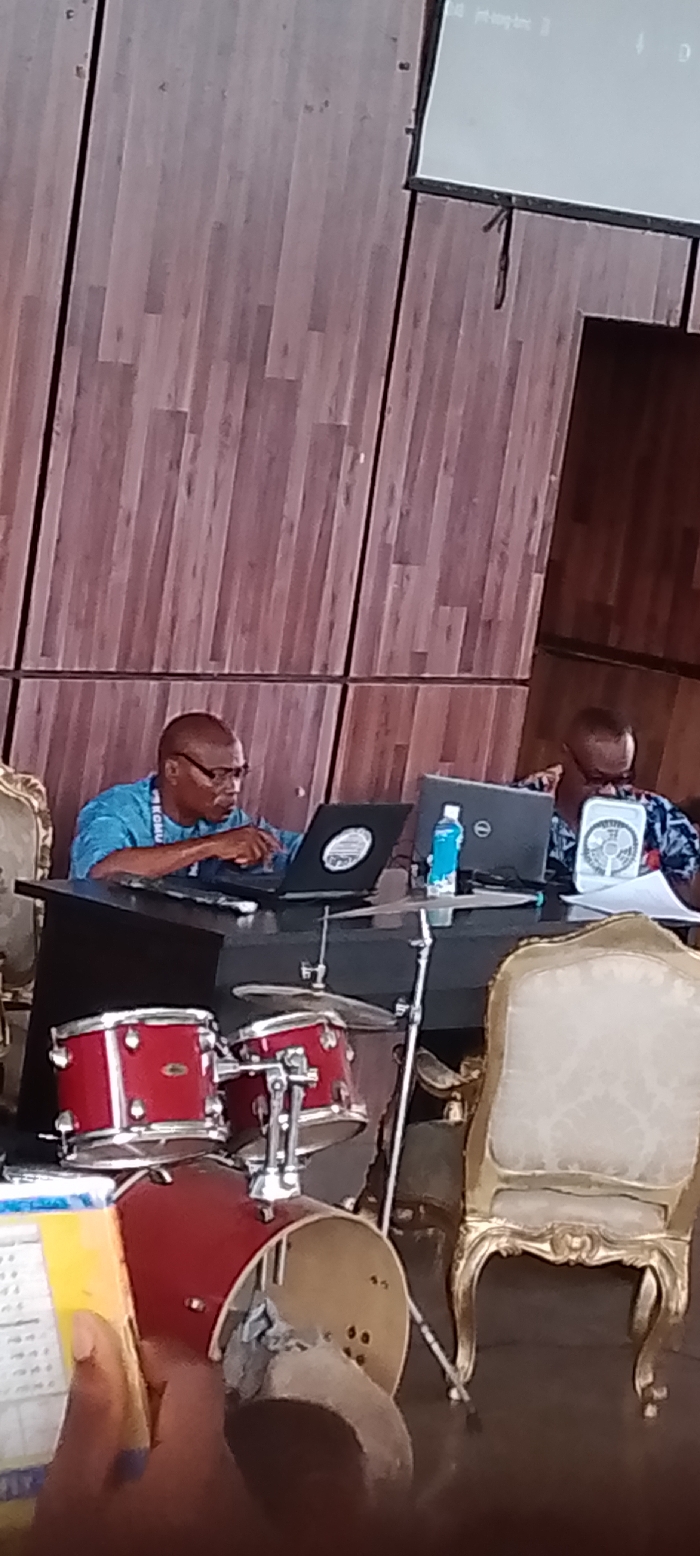 The scene is an indoor event in OGBOKO, Nigeria, featuring a partial stage or platform area. In the mid-ground, two men are seated at a dark, rectangular table. The man on the left wears glasses and a light blue patterned shirt with a lanyard, actively engaged with an open black laptop featuring a circular sticker on its lid. His left hand rests near the keyboard. The man on the right, partially visible, wears a dark patterned shirt and attends to an open Dell laptop. Between the two laptops, a light blue water bottle, a microphone, and a white circular USB fan are present on the table, alongside some white papers. In the foreground, positioned below the table, a red drum kit is visible, comprising a bass drum, two tom-toms, and cymbals on a stand. To the right of the drum kit, an ornate gold-framed chair with a light-colored upholstered seat is prominently displayed. Another similar chair is partially visible to the far left. The background consists of a tall wall with vertical light brown wooden paneling. In the upper right, a white projector screen displays text, partially readable as "id int.log.txt."