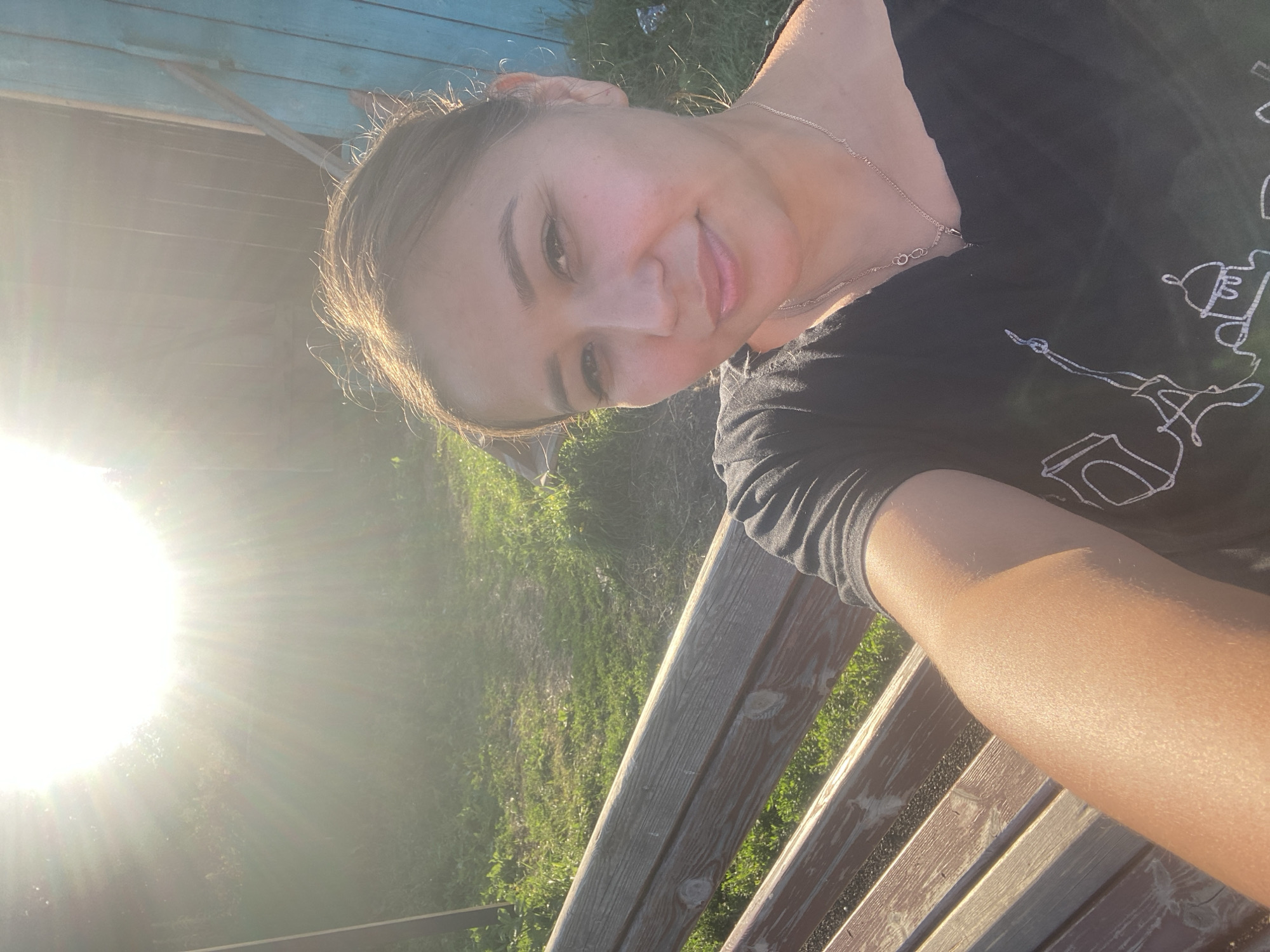 In an outdoor scene with harsh sunlight originating from the upper left, a woman with brown hair pulled back is seated on a wooden bench in what appears to be Maloarslangulovo, Russia. She is wearing a black off-the-shoulder top with a necklace. The background consists of green grass, a wooden fence painted blue on the right, and some vegetation. The bench is made of weathered wood slats. The woman is taking a selfie, with her arm and part of her leg visible in the foreground.