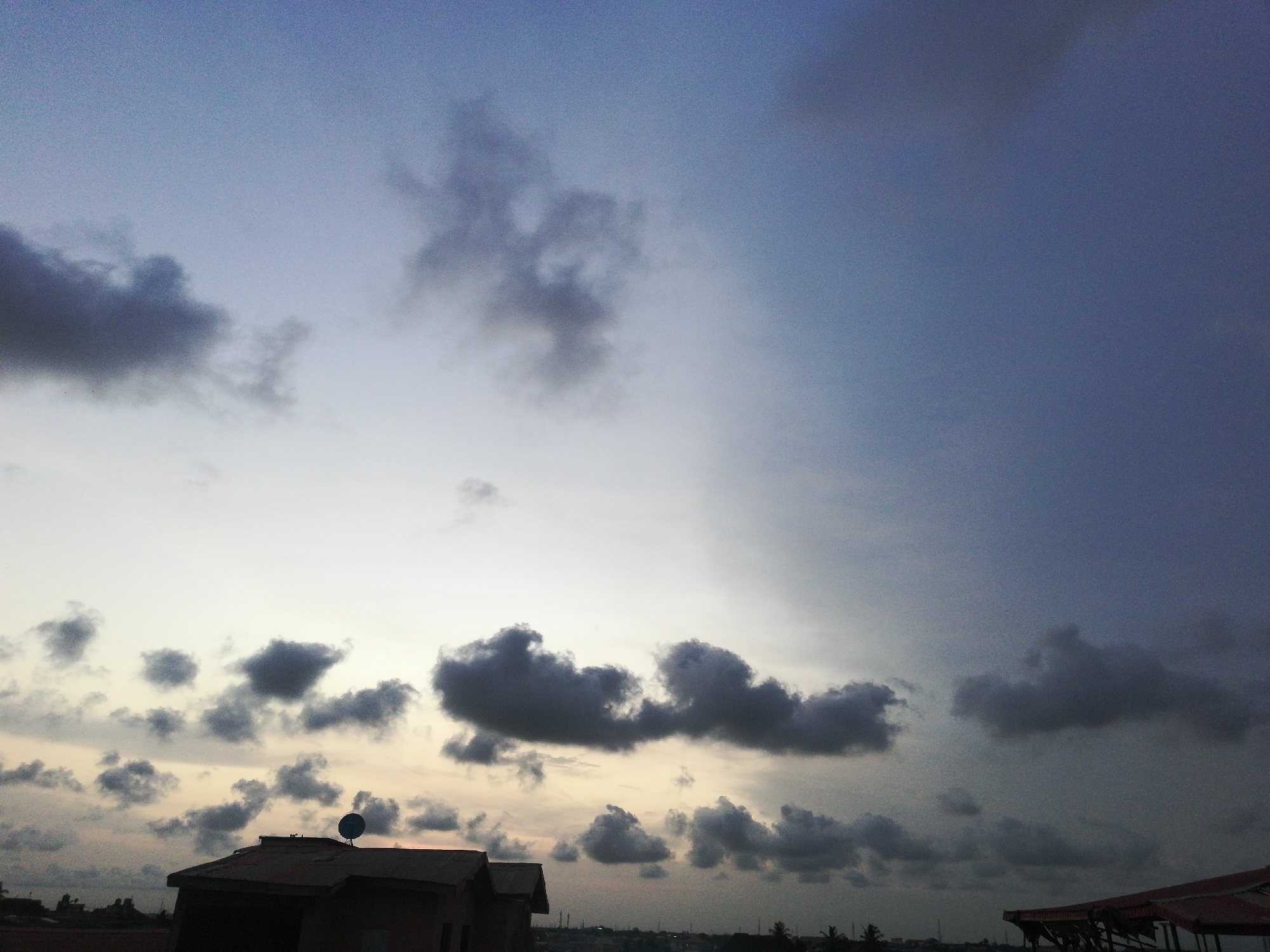 The image depicts a twilight scene in Lagos, Nigeria. A low-angle shot frames a cloudy sky dominating the view, transitioning from lighter hues near the horizon to darker shades higher up. Below the horizon line, building rooftops are visible. A house with a corrugated iron roof and a satellite dish is prominently positioned. Other buildings and vegetation are situated further in the background.