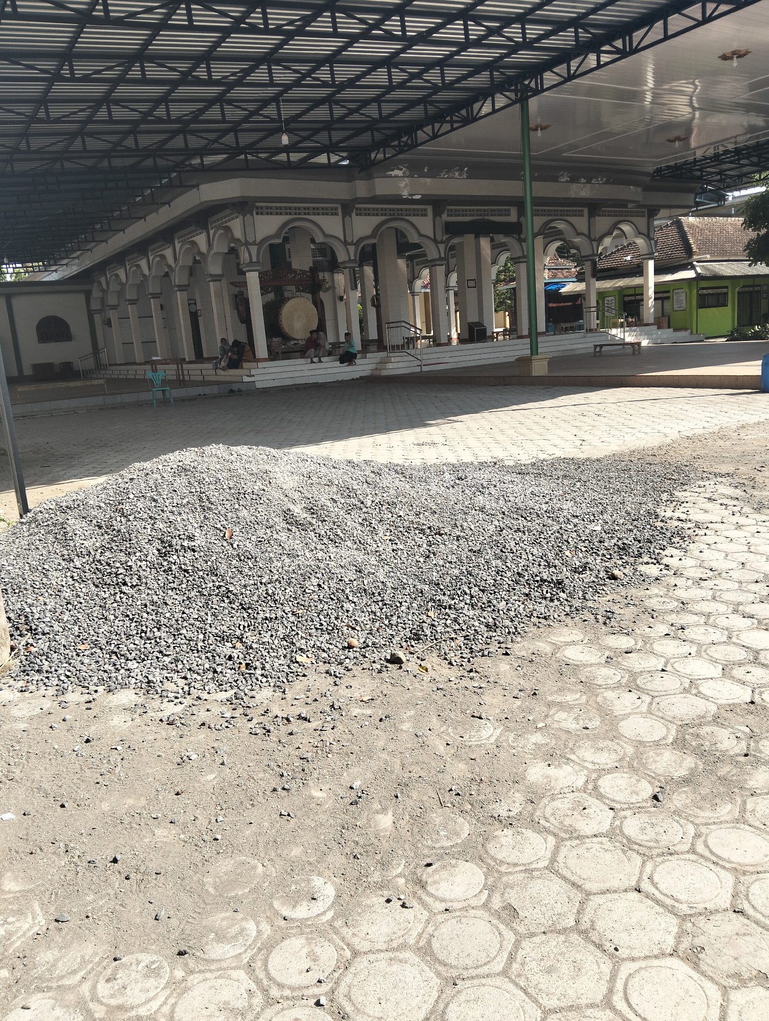 A wide-angle outdoor shot captures a sunlit scene in Wringin Putih, Indonesia, likely during daytime hours. In the foreground, a large pile of gray gravel sits on a dirt ground, extending towards the mid-ground. The gravel is composed of small, angular stones. The ground transitions from dirt to a patterned concrete surface, visible in the lower right corner. The concrete features hexagonal shapes with circular insets.
The mid-ground and background reveal a mosque complex. A wide, open roof structure with a metal frame extends over a portion of the mosque and the area in front of it, providing shade. The mosque itself has a white exterior with arched doorways and decorative elements. A large, circular drum, likely a bedug, is visible inside the mosque, mounted on a wooden frame. Several people are seated or standing near the mosque entrance, suggesting activity within the religious site. Two individuals are seated on steps leading to the mosque. A light blue plastic chair is also present.
Further in the background, a green building with a tiled roof is visible, typical of Indonesian architecture. Trees and other foliage are present, indicating a semi-urban or rural environment. The overall impression is of a community space, possibly undergoing some construction or maintenance given the presence of the gravel. The lighting suggests it is a bright, clear day.
