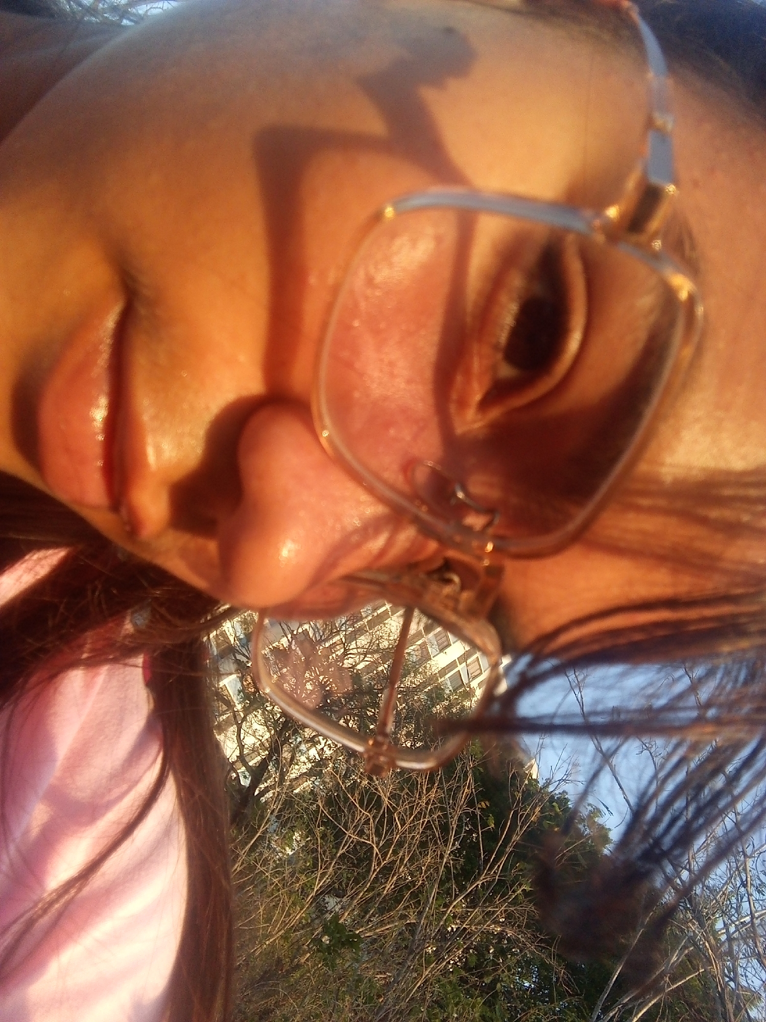 A close-up, tilted, low-angle selfie of a person with light brown skin wearing gold-framed glasses. The background, visible through the glasses and in the lower portion of the frame, shows trees with bare branches, some green foliage, a building with multiple windows, and a patch of blue sky. The person is wearing a pink shirt. It is impossible to confirm the location is Juan Griego, Venezuela, without additional information.