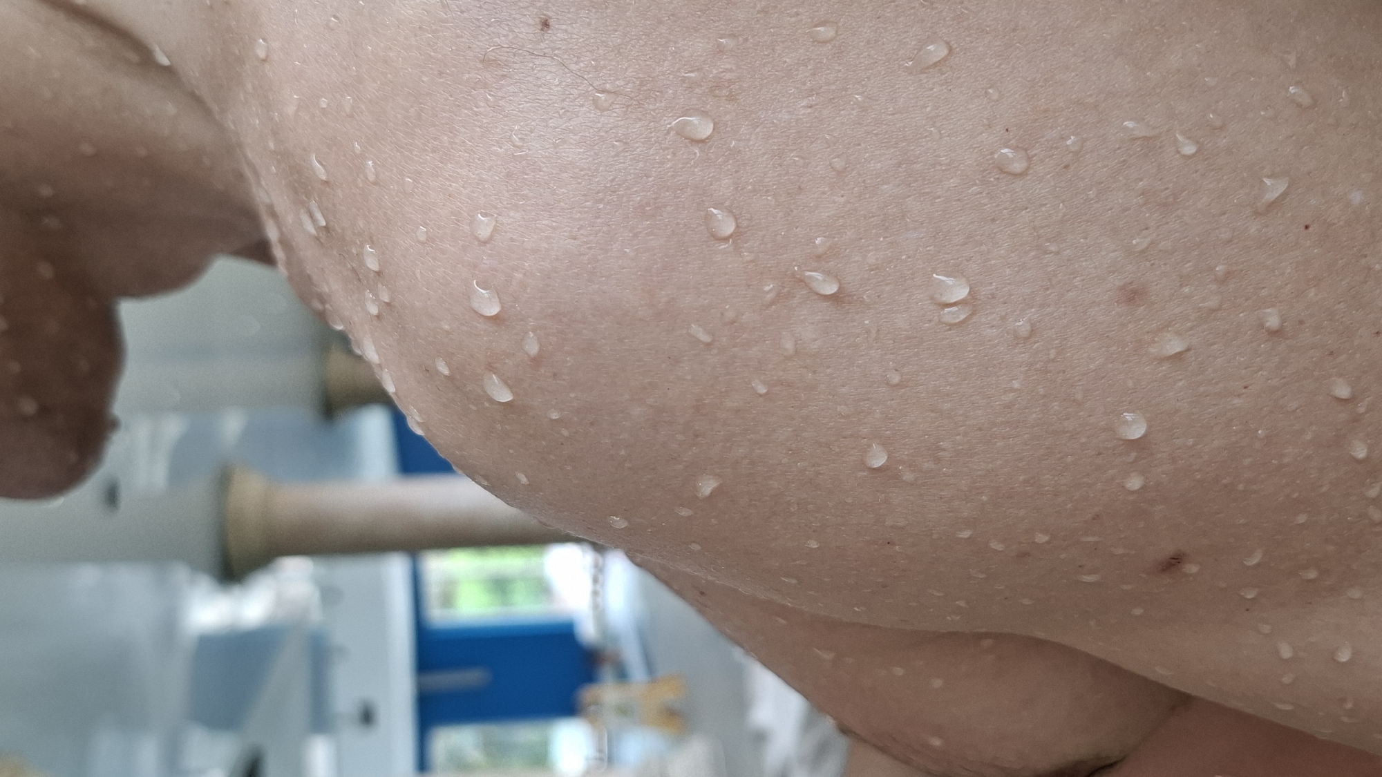 The image shows a close-up of human skin covered in water droplets. The skin is light-toned with visible pores and a few small moles. The droplets are clear and vary in size and shape. In the blurred background, there appears to be a white and blue structure, possibly a machine or building. The location is implied to be in the city of Boreti, Montenegro, but there is no explicit confirmation of the location in the visible portion of the image.