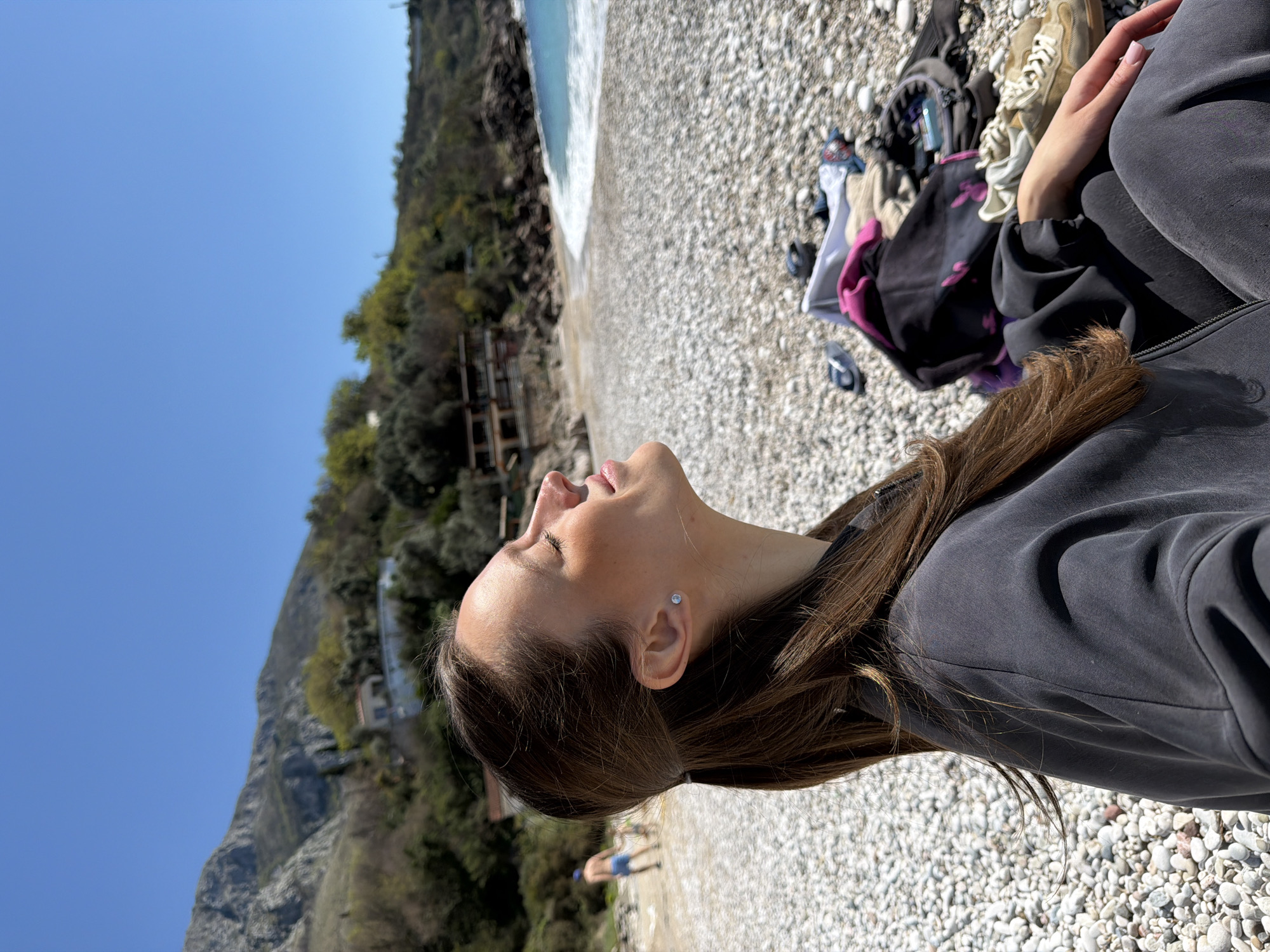 The image shows a woman lying on a pebble beach in Drobnići, Montenegro. She is wearing a gray jacket and facing upward. The beach has pebbles and some personal belongings. Beyond the beach is a body of water meeting the shoreline, and along the coast is a mountain covered in vegetation and some buildings. The sky is clear and blue.