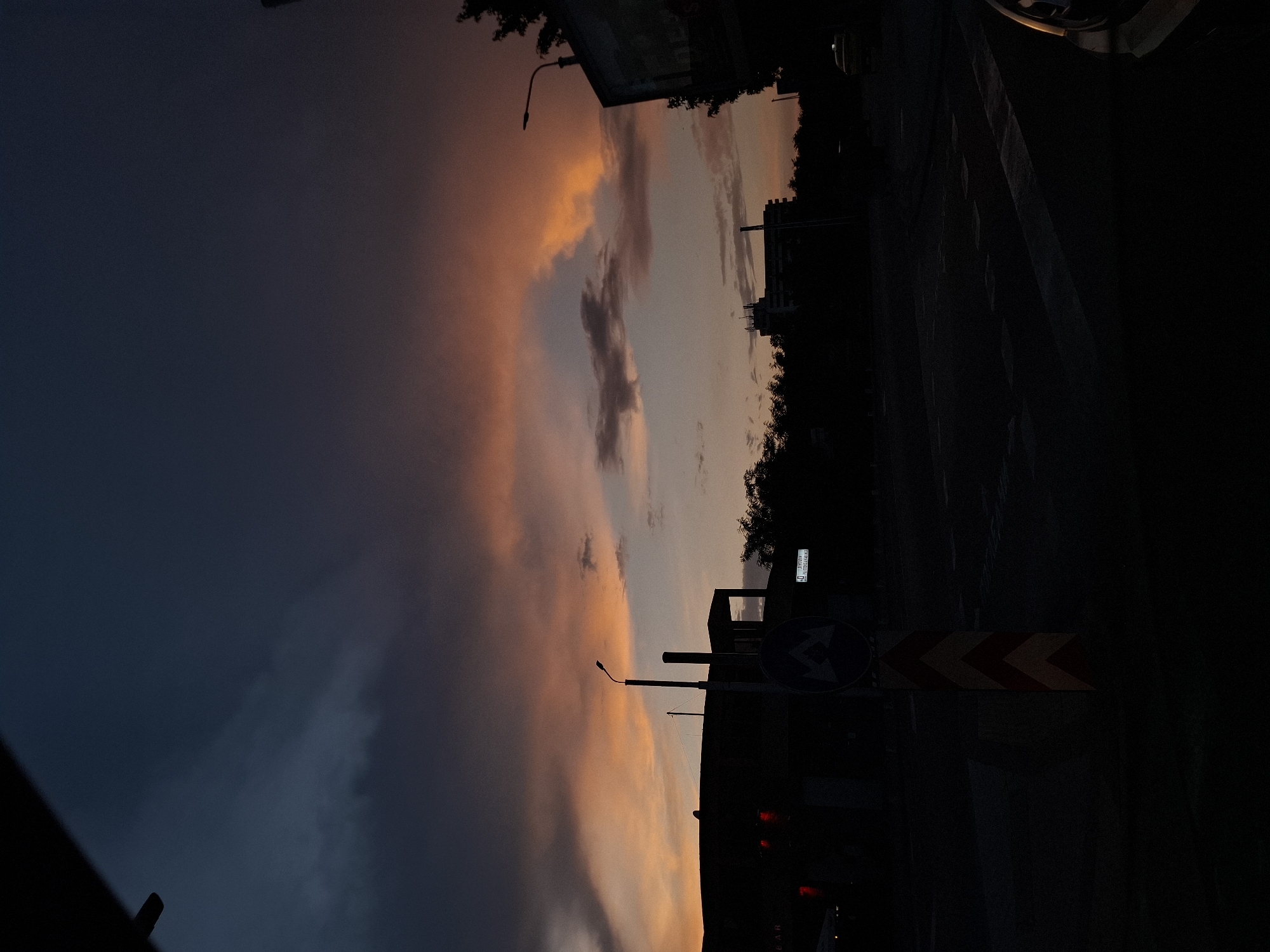 An eye-level, medium shot captures a street scene at dusk in an unidentified urban location. The sky is dominated by dramatic clouds tinged with orange and pink hues against a deep blue and dark gray expanse, suggesting the setting sun. In the foreground, a dark, silhouetted building with visible red lights is on the left, and a street sign with a double-arrow symbol is in the center. A lamppost stands tall beside it. Trees line the mid-ground, with several silhouetted buildings and structures visible behind them, including a tall, multi-story building with antennas on its roof. The road is dark and appears to be a crossroads. A sliver of a yellow car is visible in the lower right corner. The overall impression is one of a quiet, transitional moment at the end of the day.