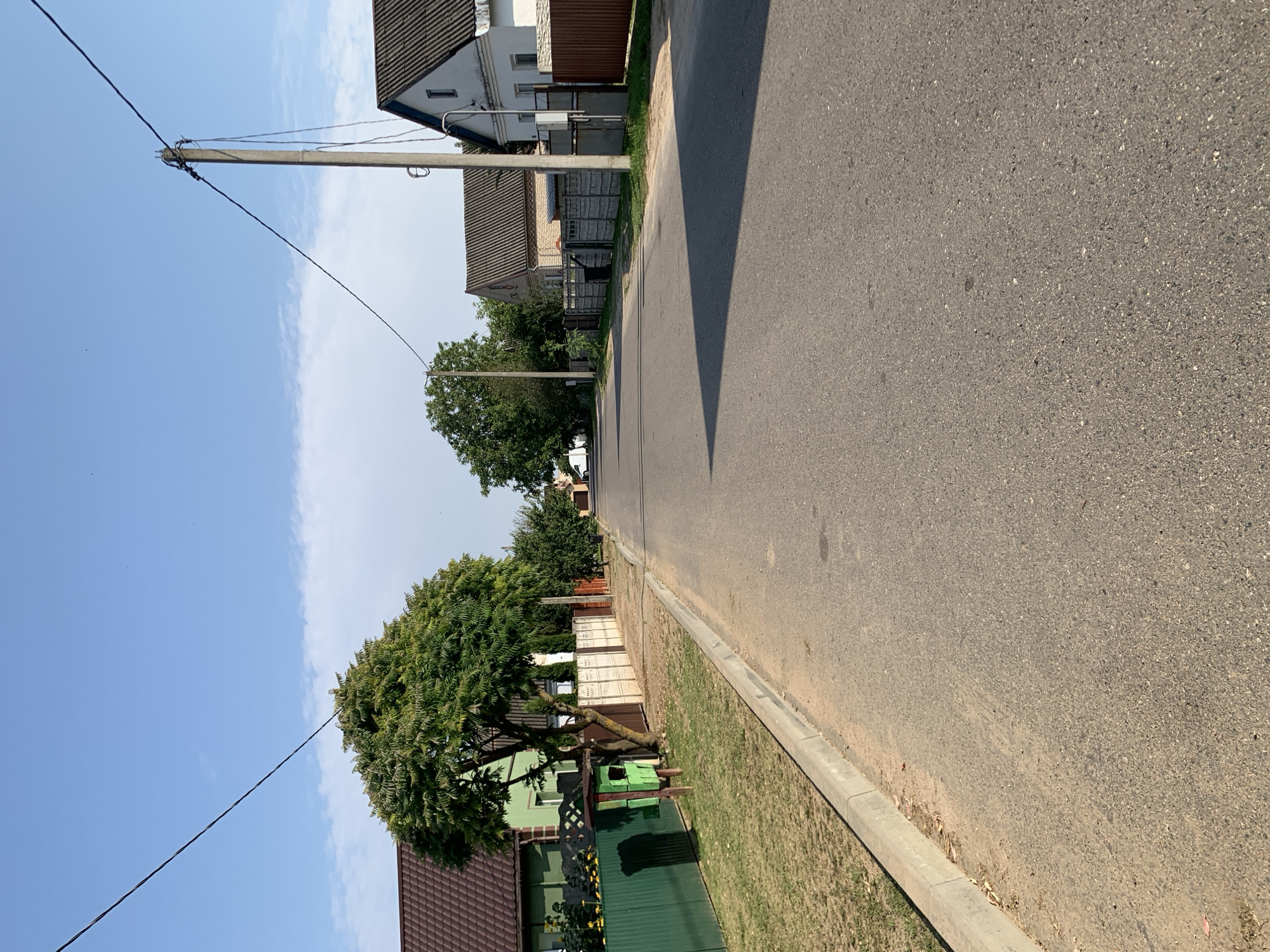 The image depicts a straight asphalt road running through a residential area in Kapyĺ, Belarus. The road is flanked by a concrete curb and a grassy verge on the left, and a sidewalk and fences on the right. Several houses with tiled roofs are visible on both sides of the road. A large tree stands on the left side, with a green wooden structure resembling a bird feeder or information booth beneath it. Overhead, power lines stretch across the blue sky, supported by a concrete pole on the right. The scene appears to be a quiet, daytime suburban street.