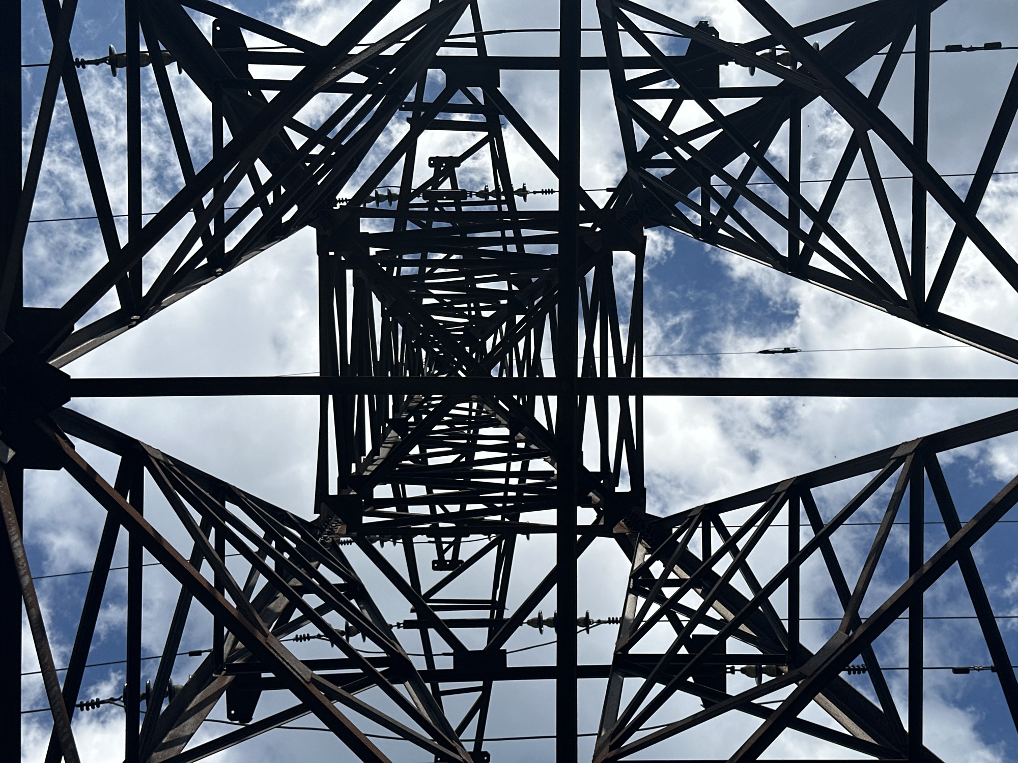 An abstract, low-angle shot captures the skeletal structure of a tall, dark metal pylon against a sky filled with scattered white clouds and a bright blue expanse. The pylon's intricate lattice of beams, supports, and wires forms a geometric pattern that dominates the frame. The metallic construction is a dark, rusty brown, appearing dense and industrial. Suspended within the structure are several ceramic insulators, each a light tan color with a ribbed surface. These insulators hang from wires, adding a touch of detail to the otherwise uniform metallic expanse. The perspective from below emphasizes the vastness of the sky and the towering nature of the pylon. The lighting suggests it is daytime, with the sky providing the primary illumination.
