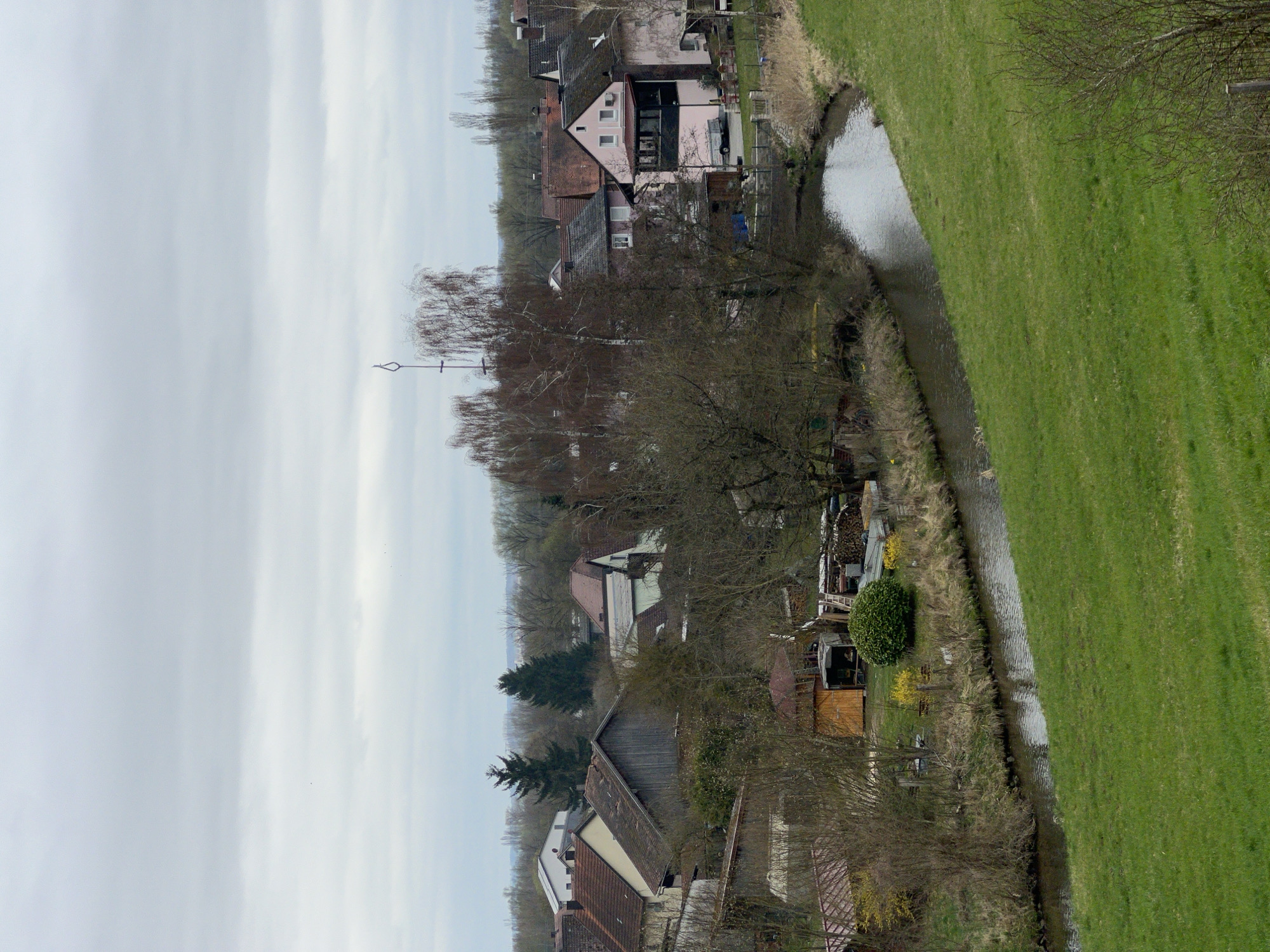 The image depicts a landscape scene in Donaustauf, Germany. On the right, a vibrant green field slopes down to a narrow stream or canal that meanders through the scene. Adjacent to the stream, there's a row of houses and buildings interspersed with trees. These buildings vary in color and design, with some appearing to have pink facades and others with more traditional roofing. The trees along the stream's edge are mostly bare-branched, suggesting a season when foliage is sparse. In the distance, more houses and buildings are visible, gradually fading into the background. Above, the sky is overcast with a layer of clouds, giving the scene a muted, diffused lighting.