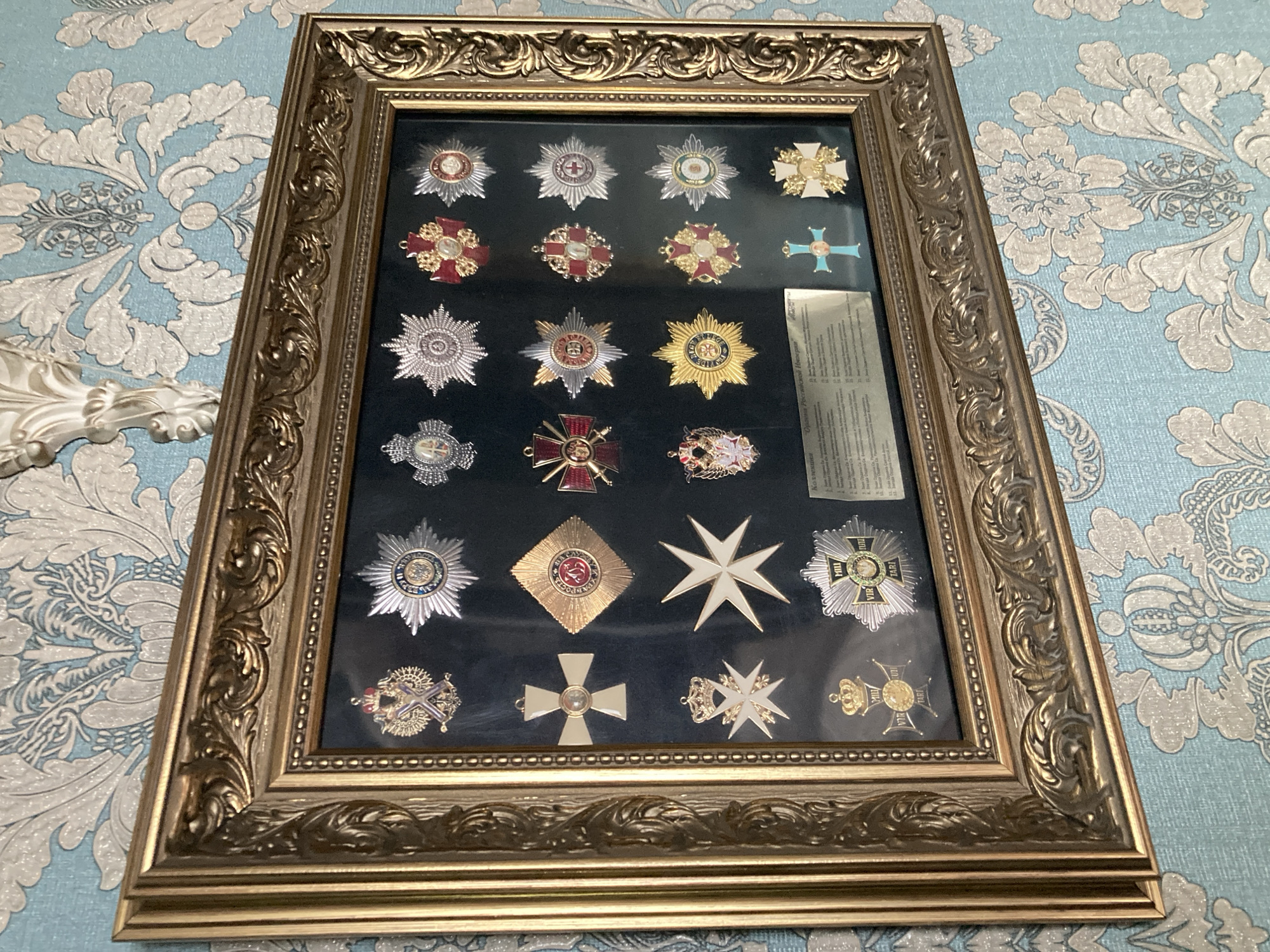 A framed collection of medals and decorations hangs on a light blue damask wallpapered wall. The frame is ornate and gold-colored. The decorations are displayed on a black background within the frame and appear to be part of a collection labeled "Коллекция Ордена Российской Империи" (Collection of Orders of the Russian Empire). The decorations are arranged in rows and columns, with various shapes and metallic finishes, including gold, silver, and enamel. Some of the medals feature intricate designs with crowns, crosses, and circular emblems. A small printed label is visible at the bottom of the display. The lighting suggests an indoor setting.