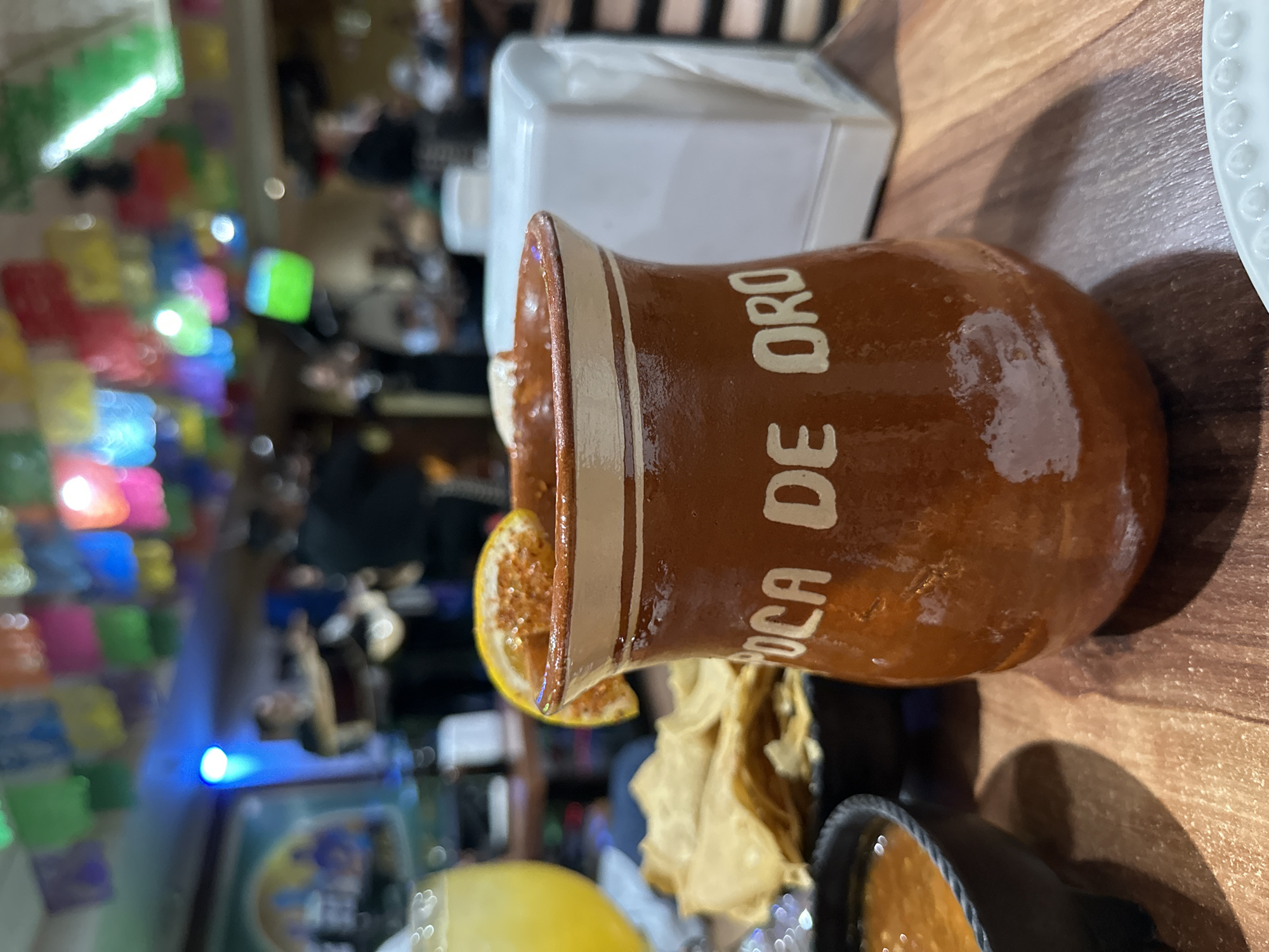 A cantaro cup with "Boca de Oro" on it rests on a wooden table in Guadalajara, Mexico. The cup is brown with white stripes and lettering. It holds a drink, garnished with a slice of orange. Next to it is a white dispenser, chips, and a bowl of salsa. The background shows a restaurant scene with colorful decorations and blurred figures of people.