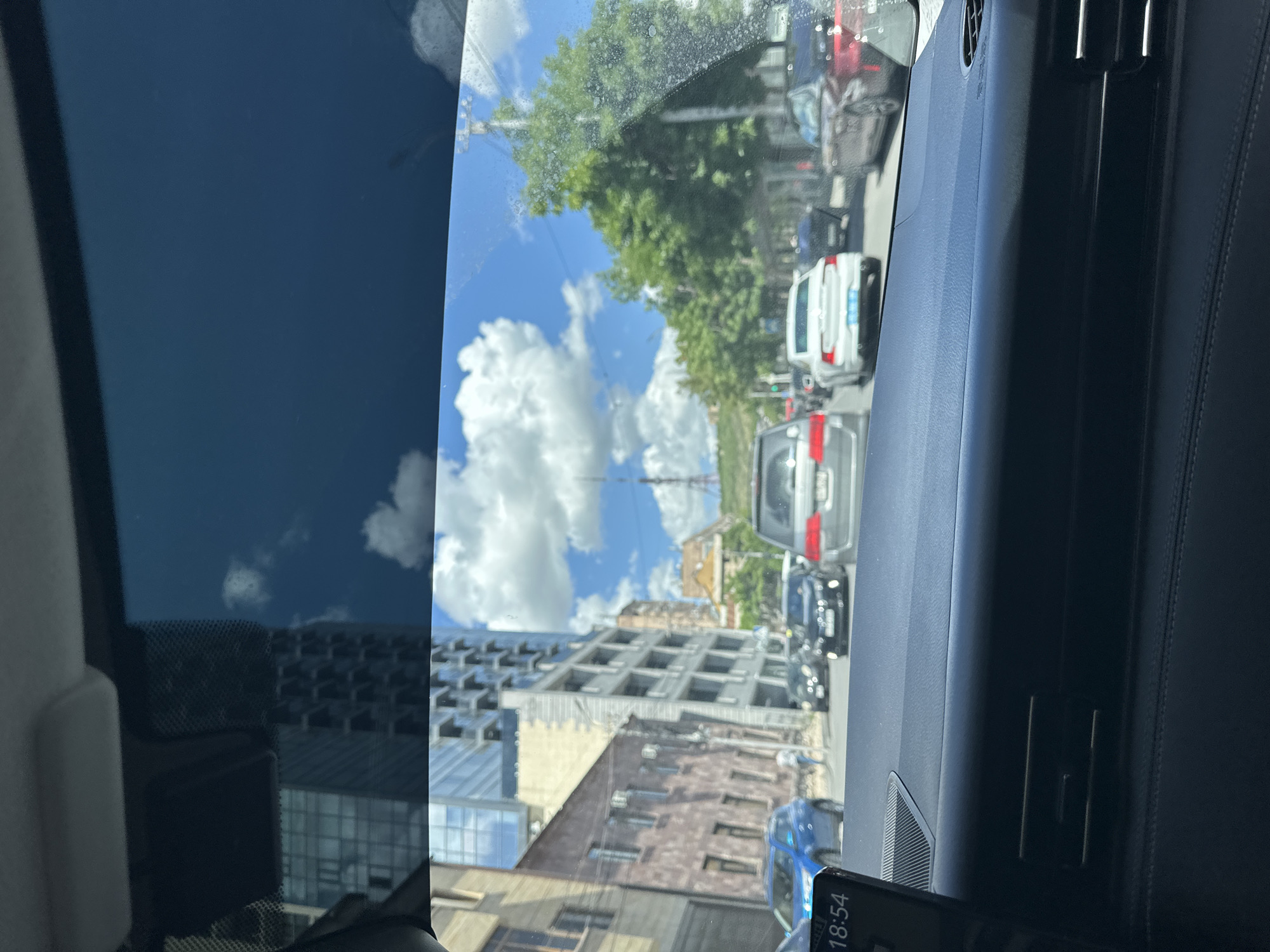 The image, taken from inside a vehicle, captures a street scene in Yerevan, Armenia, on a sunny day with some clouds in the sky. The camera is positioned behind the windshield, partially obscuring the view. The urban environment is characterized by a mix of old and new buildings, including a large, unfinished or partially constructed modern building with a grid-like facade, juxtaposed with older, traditional Armenian architecture.
Several vehicles are visible on the street, indicating moderate traffic. These include cars of various colors and models, suggesting a diverse fleet. The presence of traffic lights further reinforces the setting as an active urban street.
The overall impression is one of Yerevan as a city undergoing development, with new construction alongside existing historical structures. The image does not contain any visible people.