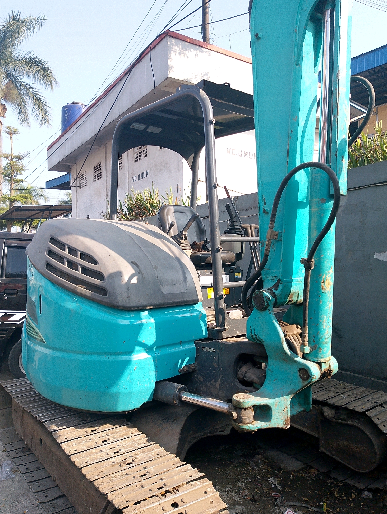 The image shows a teal-colored excavator parked on a dirt and debris-covered surface, likely outdoors in an urban or semi-urban environment. The excavator is the primary subject, with its tracks and front portion prominently visible. In the background, a white building with "WC. UMUM" (public toilet in Indonesian) signage is visible, along with electrical wires and a palm tree. The scene appears to be in Lembang, Indonesia, as indicated by the building signage. The lighting suggests it is daytime, with sunlight casting shadows. No people are visible in the image. The overall impression is one of a construction or maintenance site.