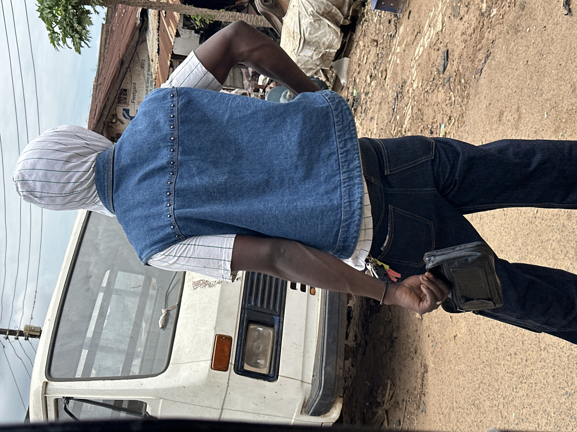 A person, viewed from behind, stands in front of a white van in a dusty urban environment in Jos, Nigeria. The person wears a white hooded garment with green pinstripes, a blue denim vest adorned with silver studs, and dark denim jeans. They hold a black wallet in their right hand. The van, with a visible headlight and grille, appears weathered. In the background, a building with a rusty corrugated iron roof and a palm tree are visible, along with power lines overhead. The ground is covered in dirt and debris.