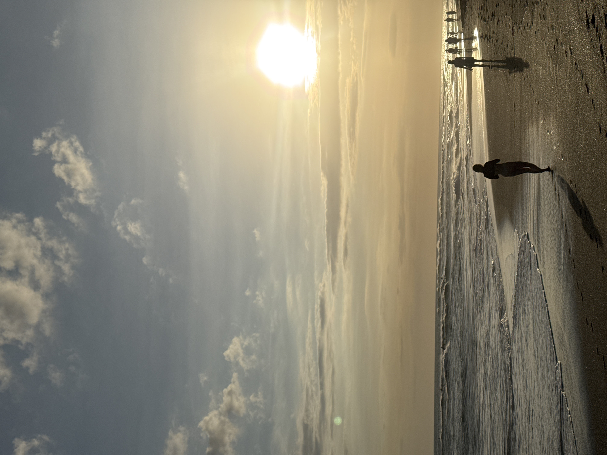 This media file depicts a serene beach scene at sunset in North Kuta, Indonesia. The sun, a bright orb in the sky, casts a warm, golden glow across the horizon, illuminating the scattered clouds and creating a dramatic sky. The gentle waves of the ocean lap against the shore, their rhythmic movement captured in the image.
In the foreground, a solitary figure stands silhouetted against the receding tide, their form etched against the wet sand. Further down the beach, a group of people walk along the shoreline, their shadows stretching out behind them, suggesting a peaceful stroll during the fading daylight. The overall mood is tranquil and contemplative, inviting viewers to immerse themselves in the beauty of the natural landscape. The image is devoid of any visible text.