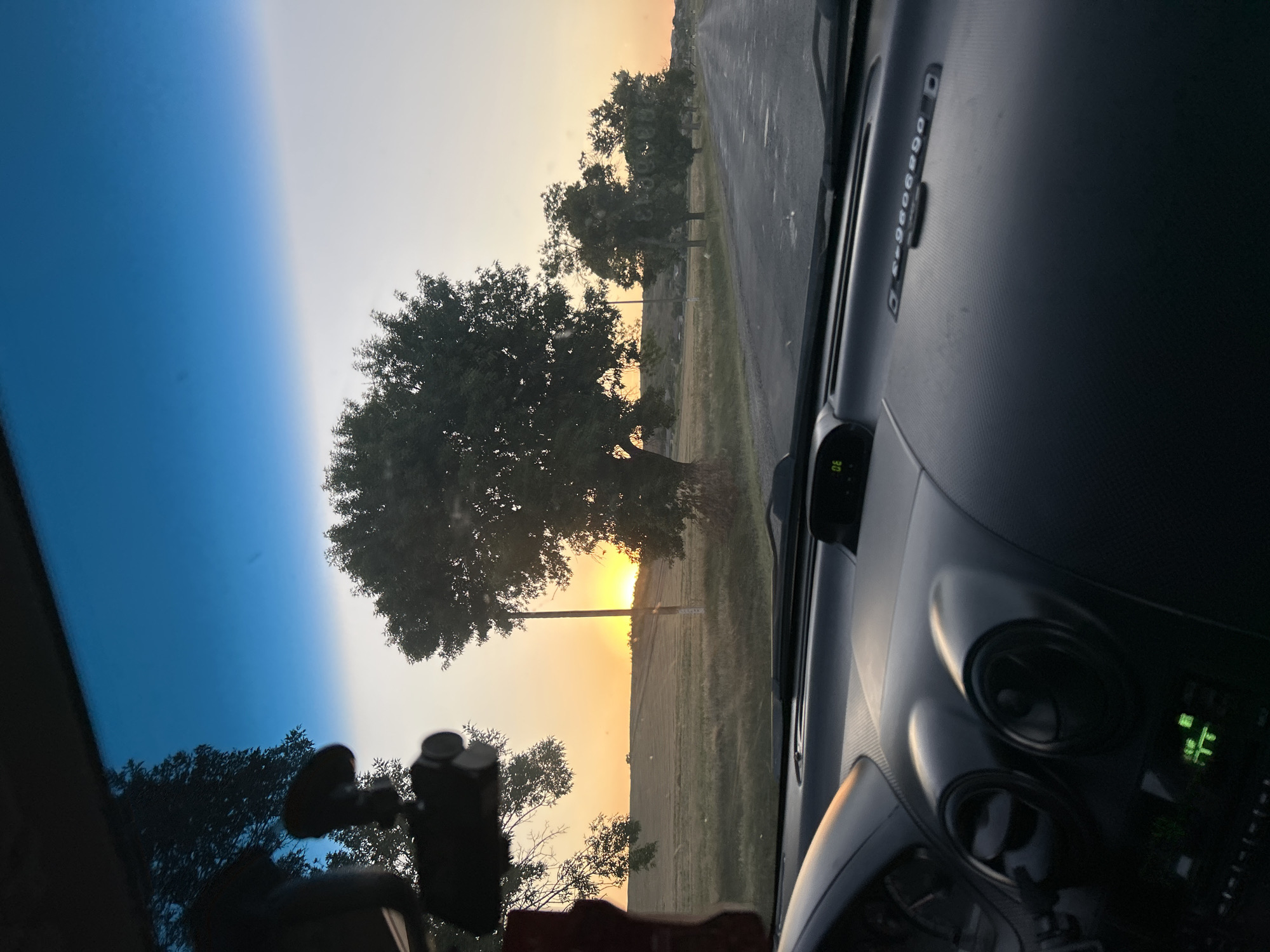 The image is a first-person perspective from inside a car, looking out through the windshield at a rural landscape during sunset. The dashboard of the car is visible in the foreground, with a dashcam mounted on the windshield. Outside, a road stretches into the distance, lined with trees on either side. The sun is setting on the horizon, casting a warm orange glow across the sky and illuminating a field. The scene depicts a car driving through the countryside of Abaclia, Moldova, at dusk.