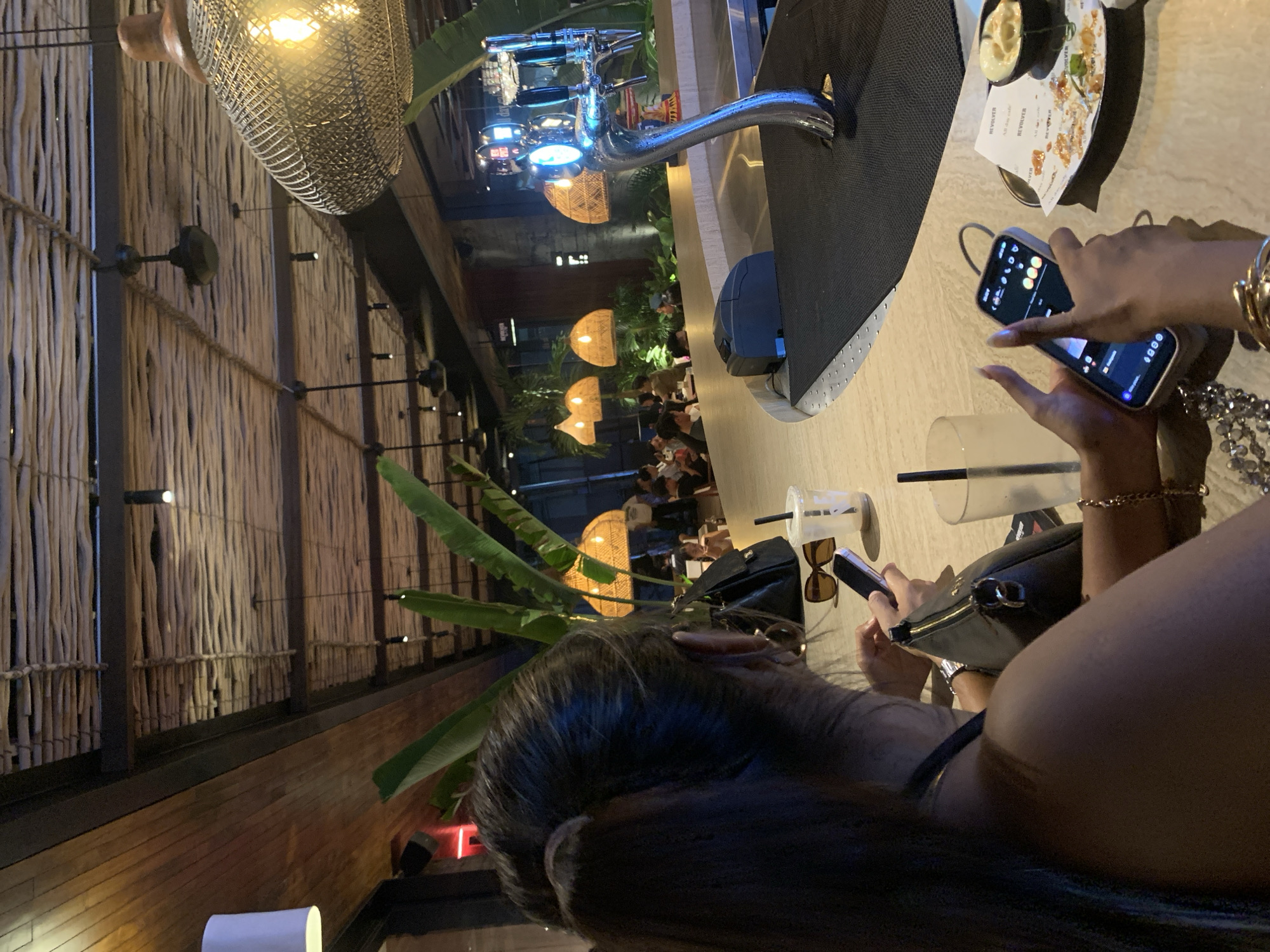 The photograph appears to be taken inside a restaurant or bar, possibly in North Kuta, Indonesia, indicated by the menu item. The image is taken from a low angle, presenting a view from the perspective of a patron sitting at a bar. The focus is on the upper body of a person with long dark hair who is looking at a mobile phone in their hands. The person has gold bracelets on their wrist. The phone's screen is illuminated and displays an image. The background of the shot features the interior of the restaurant. Overhead, there are decorative lamps. People are seated at tables and appear to be dining. The bar area includes a chrome tap system. A drink is on the bar top in front of the phone. The overall ambiance suggests an evening setting with dim lighting.