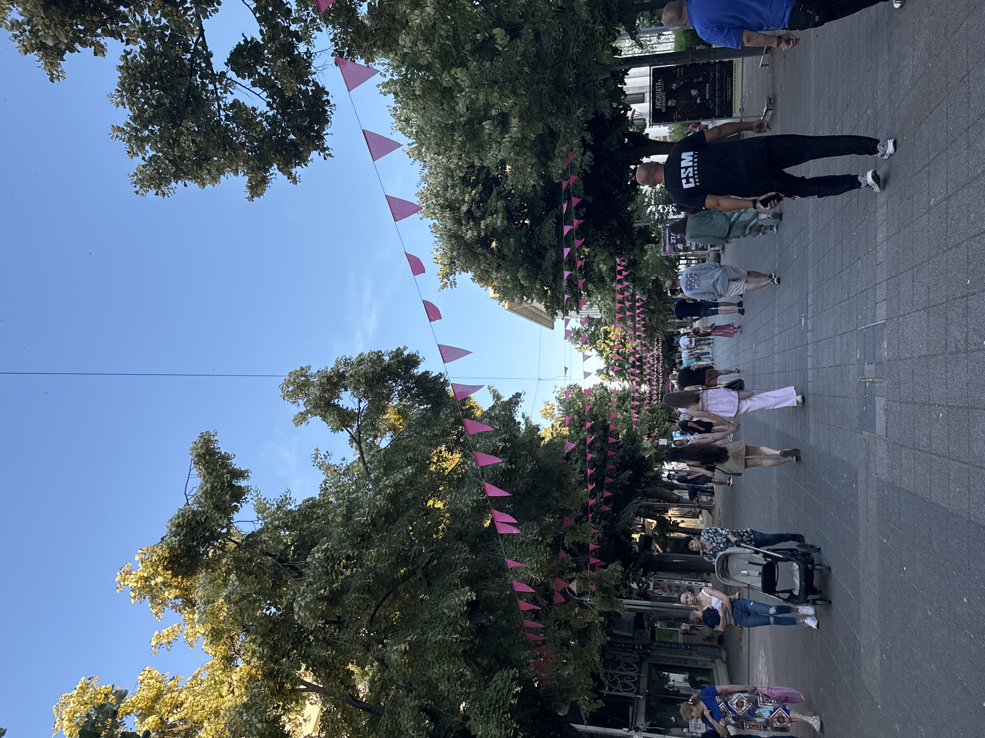 The image depicts a pedestrian street in Burgas, Bulgaria, likely during daylight hours given the clear blue sky and sunlight illuminating the trees. The street is lined with mature trees on both sides, their branches creating a canopy overhead. Pink triangular flags are strung along lines above the street, adorning the thoroughfare.
Several people are walking along the paved street, some singly, some in pairs, and a few in small groups. On the left side of the image, a woman in a patterned dress walks with a pink bag. Next to her, a young woman in ripped jeans holds a baby, and an older woman pushes a stroller. Further down the street, a group of people are walking away from the camera. One man in the foreground is wearing a black t-shirt with "CSM BUCHARESTI" printed on the back and black sweatpants. A poster advertising a performance is visible on the right side of the street. The atmosphere appears to be relaxed and casual, suggesting a public space frequented by locals and possibly tourists.