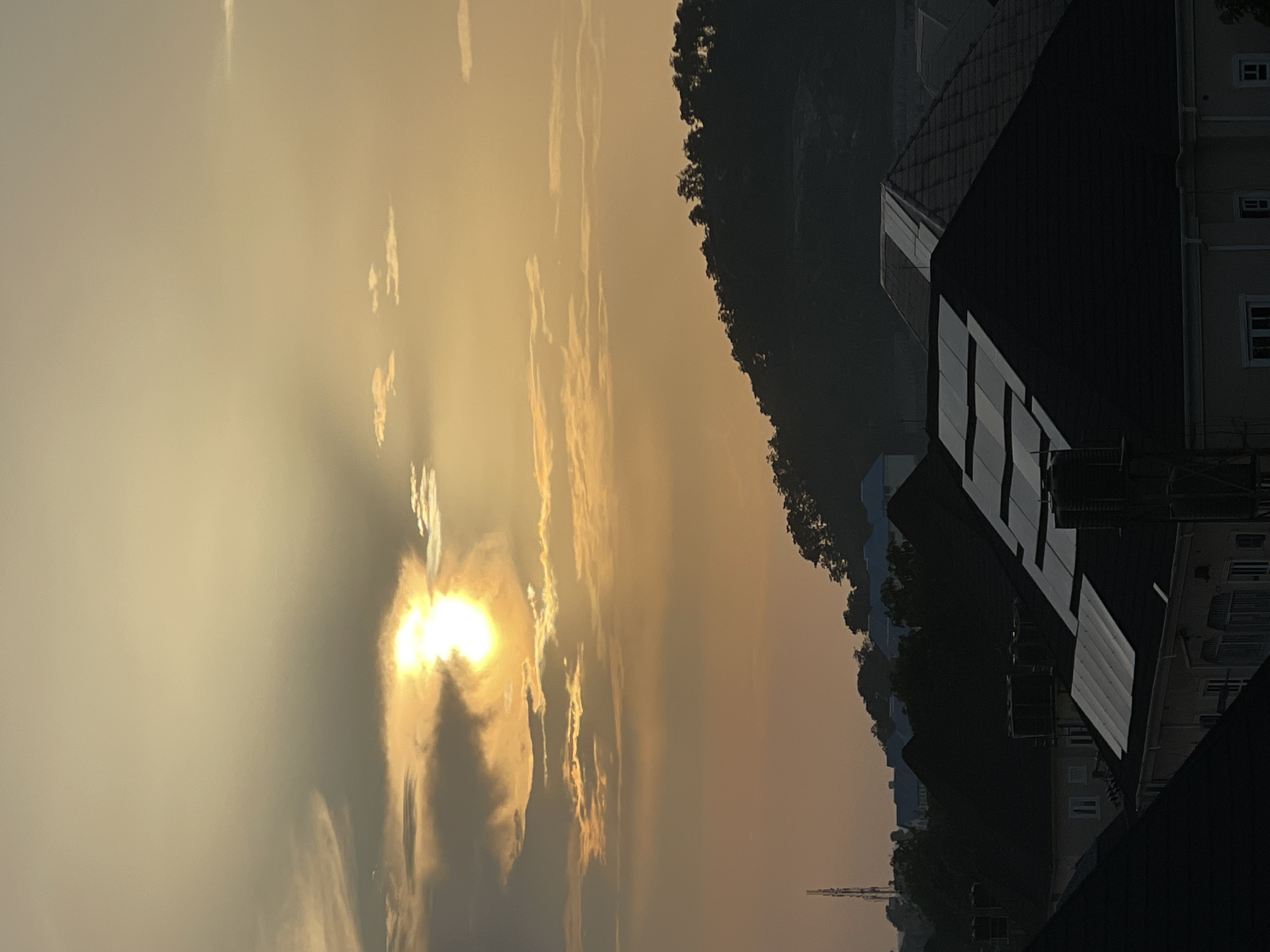 The image captures a sunset over a residential area in Abuja, Nigeria. The sky is a warm gradient of orange and yellow, with the sun partially obscured by clouds, creating a soft, diffused light. Below the sky, a dark, tree-covered hill slopes downwards, forming a silhouette against the twilight. In the foreground, several houses with dark roofs are visible, some with visible solar panels. The overall impression is of a serene evening scene in a suburban environment. The presence of a distant telecommunications tower suggests an urban or semi-urban setting, consistent with the location in Abuja. No people are visible in the image.