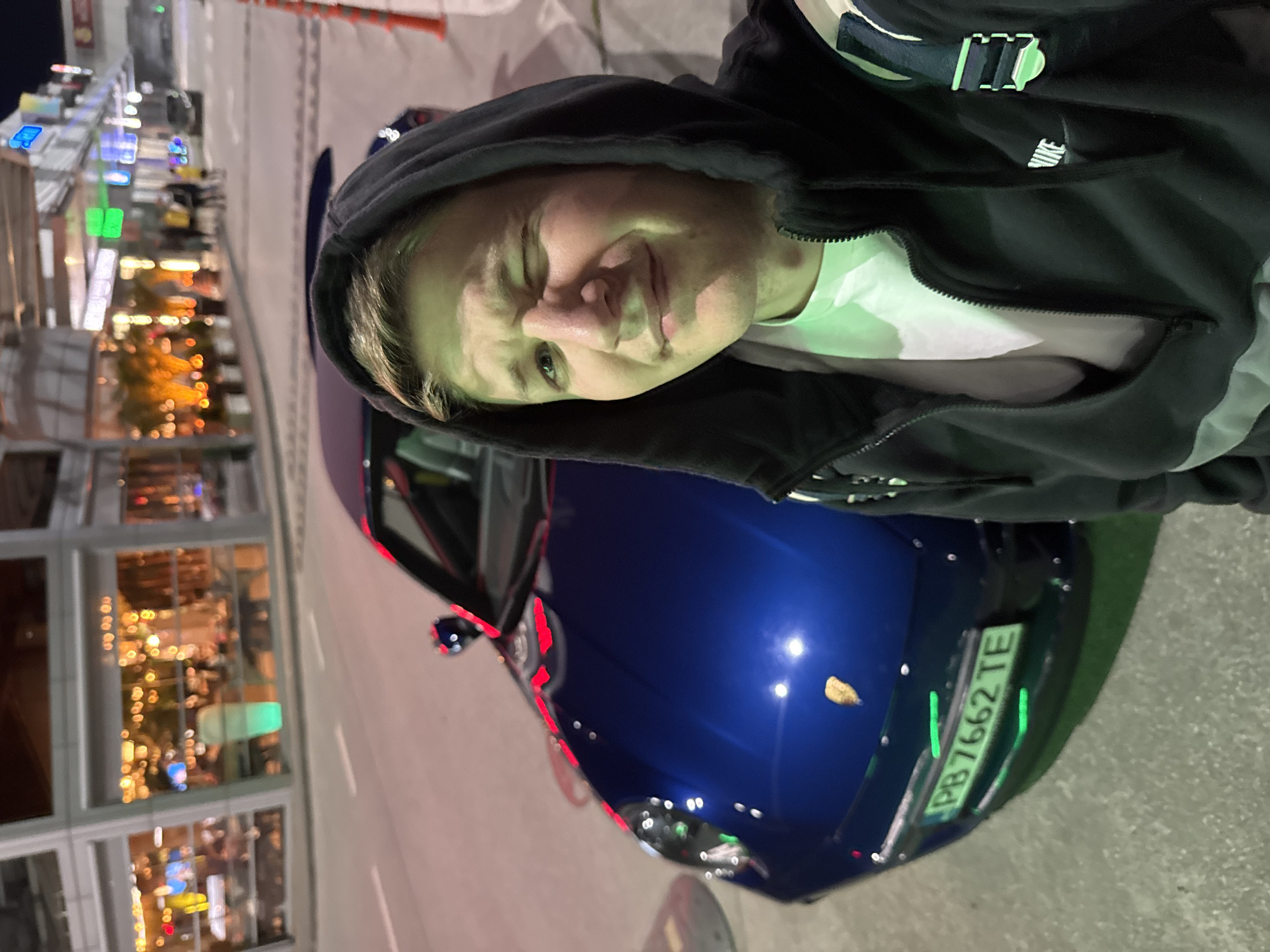 The image displays a young individual standing outdoors at night, likely in Plovdiv, Bulgaria, next to a dark blue car. The person, positioned in the foreground, is looking towards the camera with a slight, neutral expression, their face illuminated by a noticeable greenish light source. They are wearing a black hooded jacket with the hood up, over a white t-shirt. A white "Nike" logo is visible on a strap or shoulder of their jacket.
Behind and to the left of the person, a gleaming dark blue car, possibly a sports car, is partially visible, showcasing its rear quarter and trunk. The car's reflective surface catches bright artificial lights from the surroundings. A Bulgarian license plate, reading "PB7662TE," is clearly seen on the back of the vehicle, with "PB" indicating Plovdiv.
The background reveals a building with large windows reflecting numerous warm-toned lights and some distinctly illuminated green and blue signs, suggesting an urban environment with commercial establishments or storefronts. The ground is a dark, paved surface. The image is taken at night, as indicated by the prominent artificial lighting and reflections.