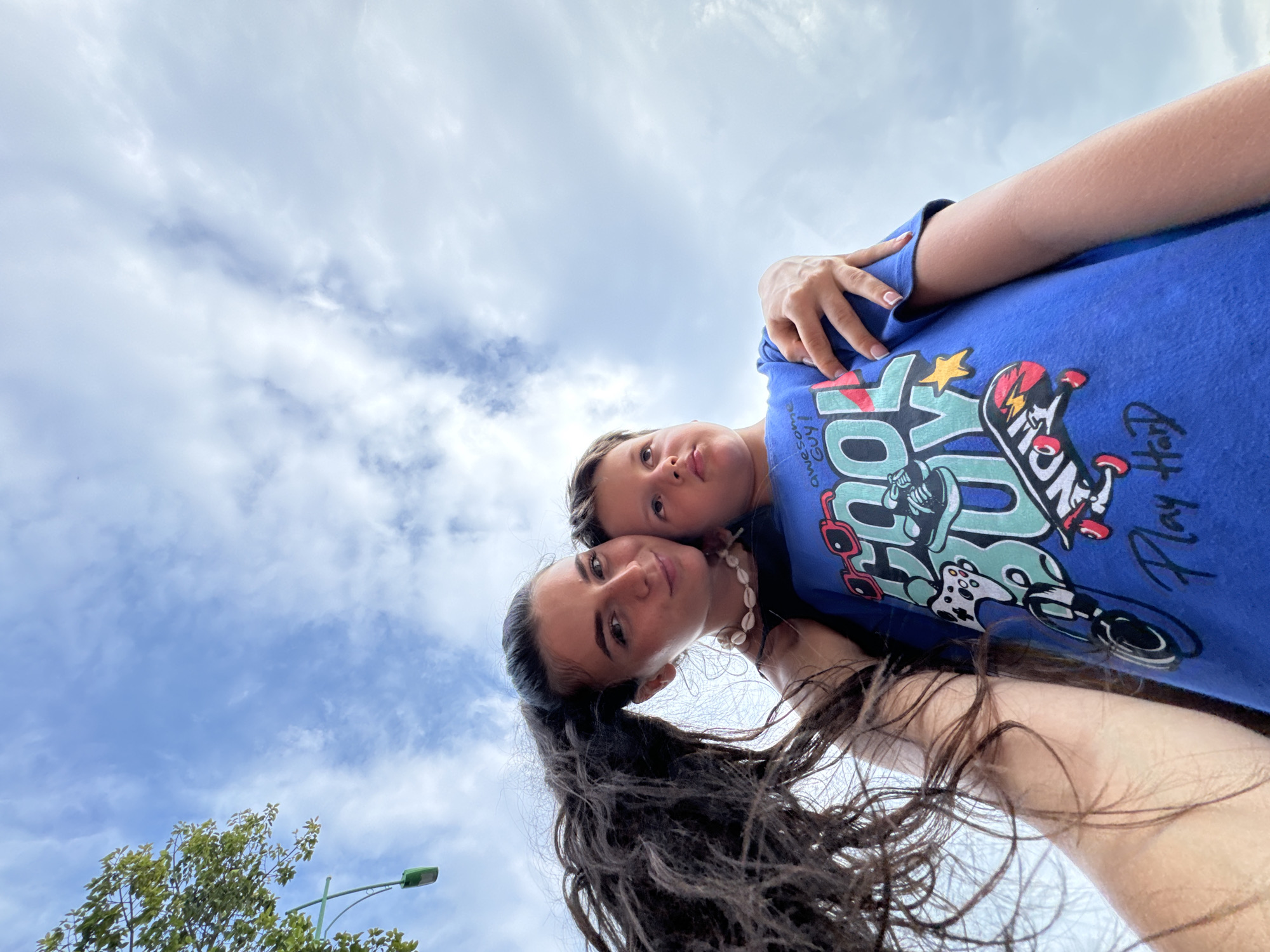 This low-angle, outdoor portrait features two individuals, an adult female and a child, captured against an expansive cloudy sky. The camera looks up at the subjects, who are positioned closely in the foreground with their heads touching.
The adult female is on the left, sporting long, dark, curly hair that cascades downwards due to the perspective. She wears a shell necklace and a dark top, looking towards the camera with a serene expression. The child, likely male, is nestled behind her, resting his head against her side. He is dressed in a vibrant blue t-shirt embellished with a colorful graphic design featuring a skateboard, controller-like shapes, and text. Visible phrases on his shirt include "awesome Guy!", "OY!", "Play Hard", and "Skate".
The background is dominated by a bright, overcast sky with scattered white and light blue clouds, suggesting daytime. In the bottom left corner, the green foliage of a tree and a green lamppost are partially visible, indicating an outdoor setting. The image conveys a moment of closeness and contentment between the two subjects, possibly captured as a selfie. The scene is set in Phan Thiết, Vietnam.