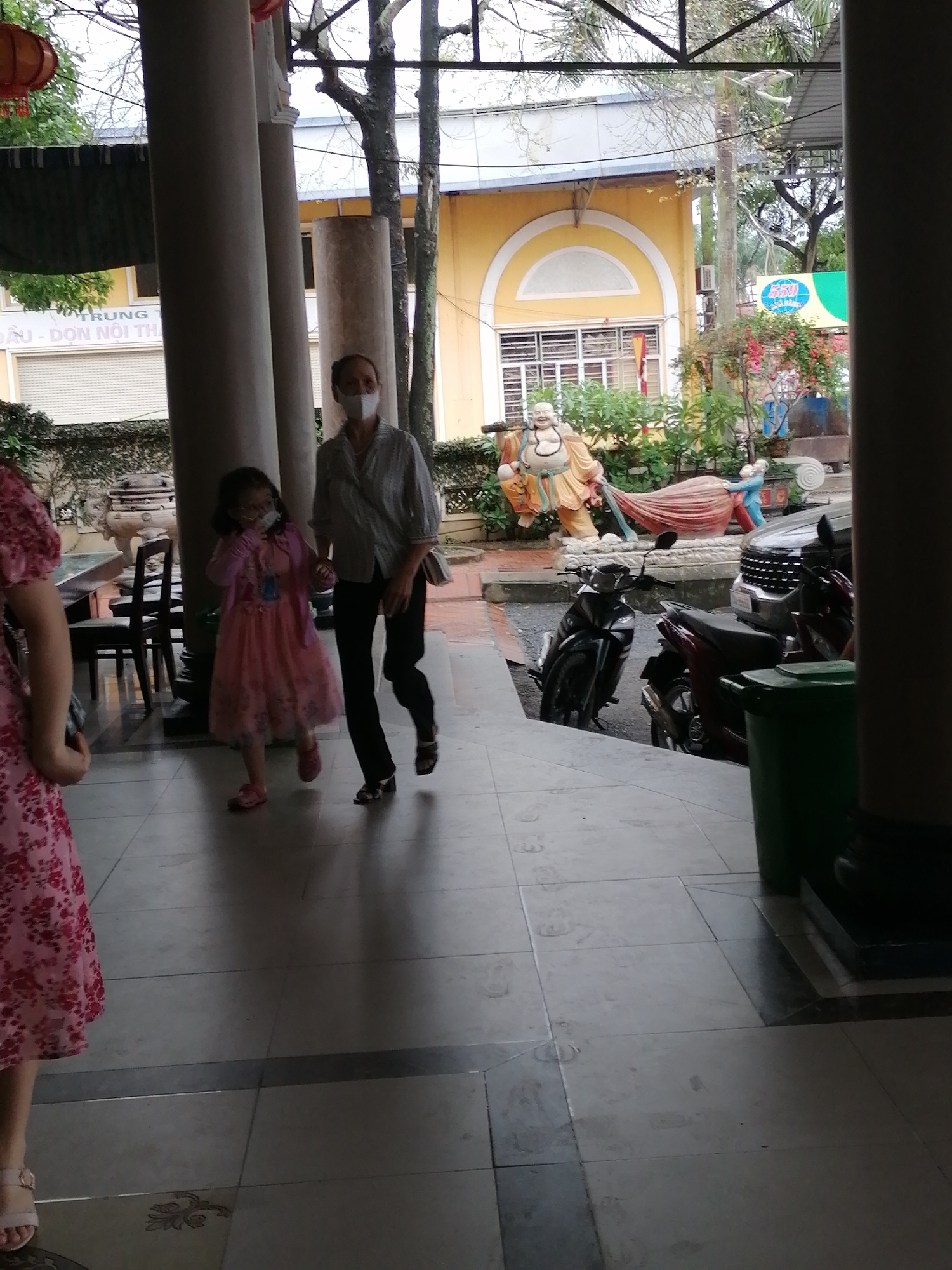 Captured from an indoor perspective in Chu Văn An Ward, Vietnam, the image shows a woman and a young girl walking towards the camera. Both are wearing face masks. The woman is in a button-down shirt and dark pants, while the girl wears a pink dress. A figure in a floral dress is partially visible in the lower left corner. Outside, various scooters and cars are visible, along with statues of a Buddha and other figures. A yellow building with arched windows stands in the background. The shot is framed by pillars, suggesting the view is taken from a covered area.