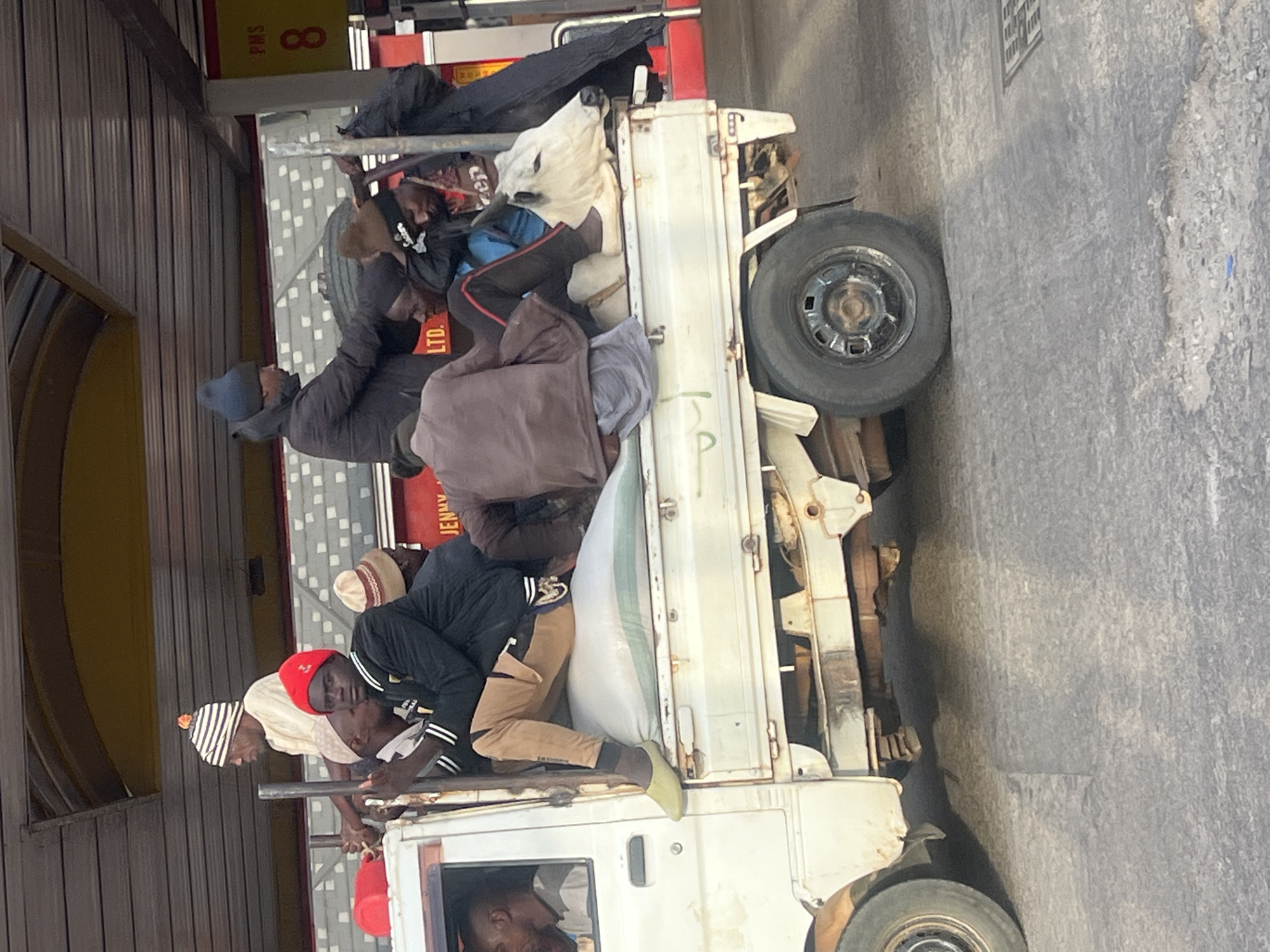 The image captures an overloaded white pickup truck, likely in Jos, Nigeria, transporting multiple men and large white sacks in its open bed during daylight hours. At least seven men are visible in the truck's cargo area, situated amongst and on top of bulging white sacks, which appear to hold bulk goods. The men are dressed in casual attire; notable headwear includes a red beanie, a striped beanie, and a dark blue beanie. Their expressions are largely neutral, suggesting a routine journey or a stoic acceptance of the conditions. One man is partially visible inside the truck's cabin.
The vehicle itself is a basic utility pickup truck, showing signs of use. In the background to the left, a building with corrugated metal walls can be seen, possibly a commercial or industrial structure. Further back, a red surface or vehicle displays partial text, including "PMS 8" and "JENNY LTD.". The ground beneath the truck is paved with asphalt or concrete. The bright, even lighting indicates it is daytime with clear weather. The scene reflects a common mode of transportation, often involving overcrowding, in certain contexts.