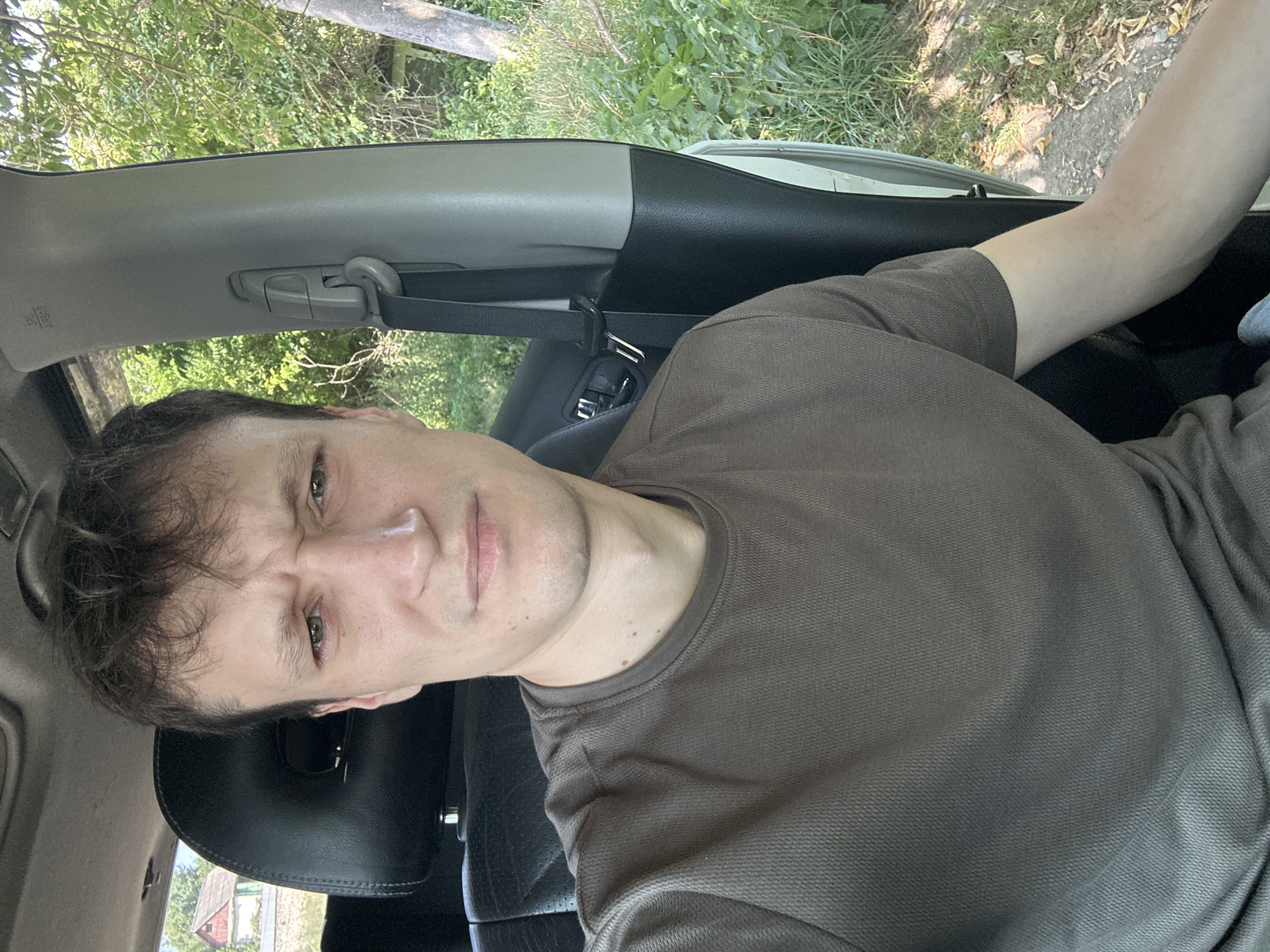 A man is sitting in the driver's seat of a car, looking at the camera with a confused expression. He is wearing a brown t-shirt. The car's interior is visible, including the black leather seat and a grey pillar with a seatbelt. Outside the car, lush green trees and foliage can be seen, suggesting a rural or suburban setting. A house with a red roof is visible in the background to the left. The lighting indicates it is daytime. The provided crops highlight close-ups of the man's face, the car's interior, his clothing, and the surrounding greenery.