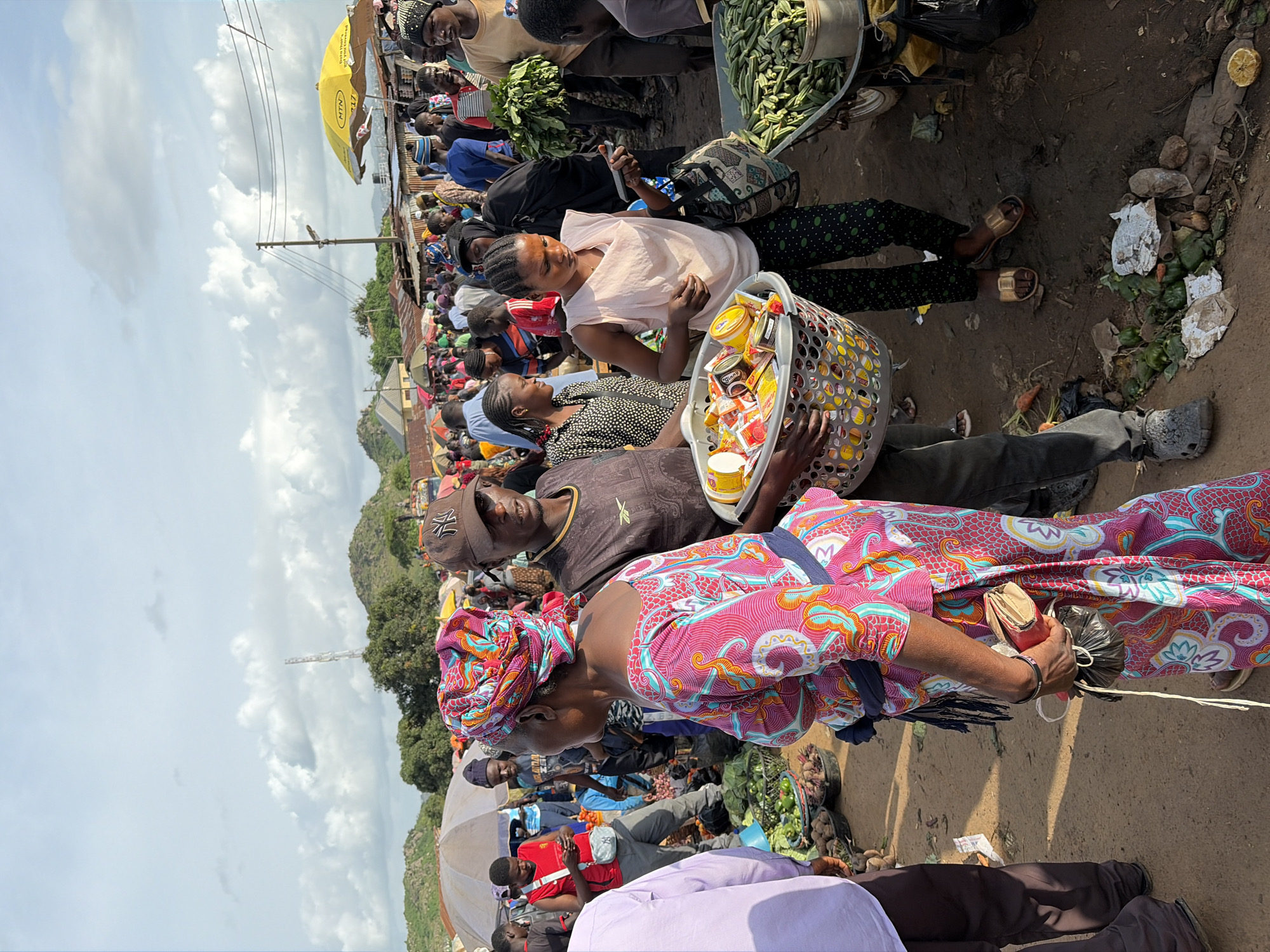 A street market scene is depicted in Gura Top, Nigeria. A diverse crowd of people, predominantly of African descent, are engaged in commerce. In the foreground, a woman in a vibrant pink and blue patterned dress and headscarf holds a red wallet and a black plastic bag. To her right, a man wearing a brown cap and dark t-shirt carries a large basket overflowing with various food items, including canned goods and what appear to be spice packets. Adjacent to him, a young woman in a light pink sleeveless top and black polka dot pants examines a mobile phone while holding a patterned tote bag. Behind them, vendors display produce like okra and root vegetables in baskets and on carts. The market is bustling with activity, with numerous stalls and umbrellas providing shade. The ground is dusty, indicative of an outdoor market. In the background, low-lying hills and a communication tower suggest the market is situated in a semi-urban or rural area. The sky is partly cloudy, with a mix of sunlight and shadow across the scene.