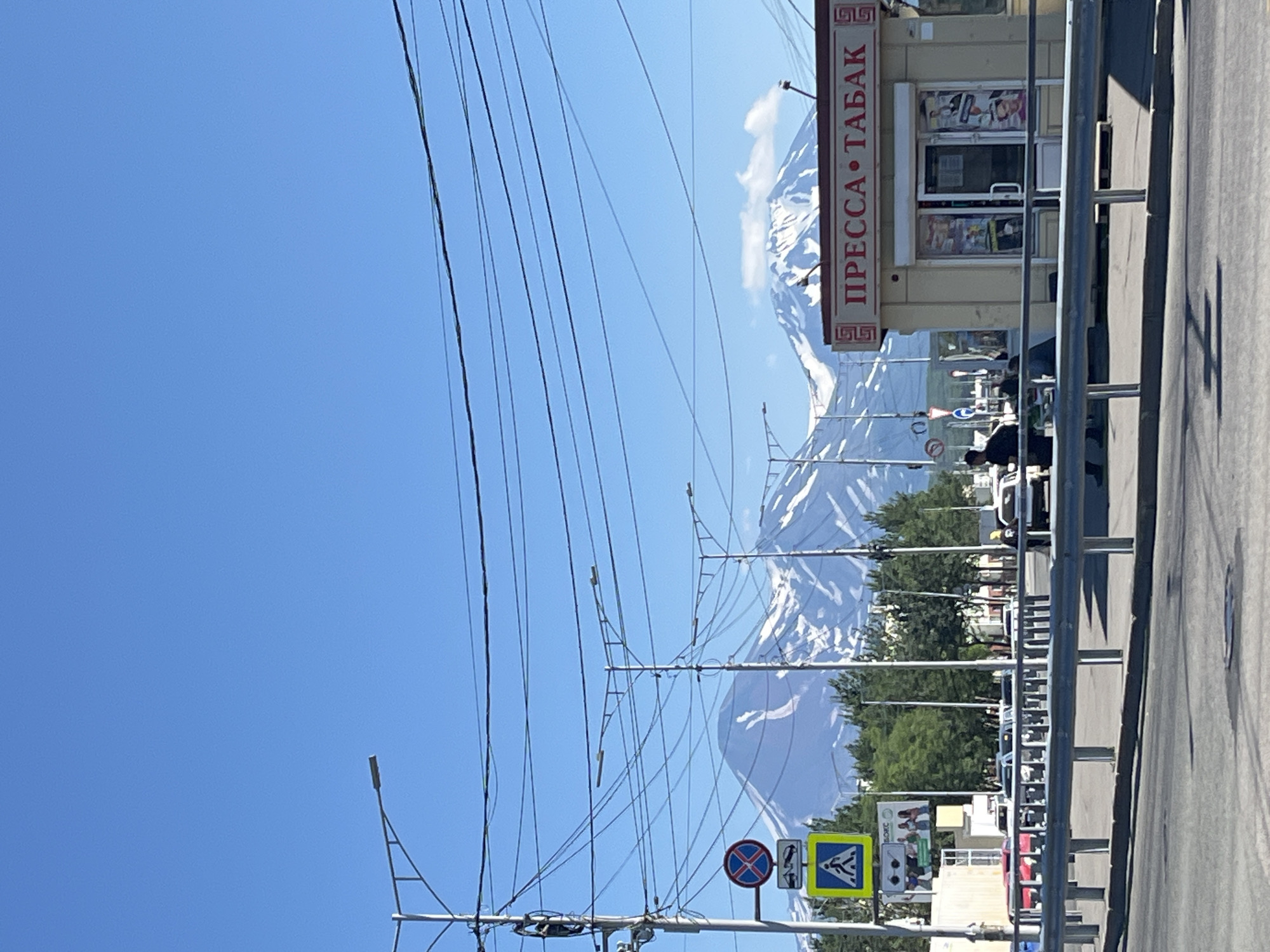 This is a street-level, medium-distance, eye-level shot of a street in Petropavlovsk-Kamchatsky, Russia, on a clear, sunny day. In the background, two snow-capped volcanoes loom under a bright blue sky, with power lines crisscrossing the scene. In the middle ground, a small kiosk with the Cyrillic text "ПРЕССА • ТАБАК" (Press • Tobacco) is visible, displaying magazines and newspapers. Several people are near the kiosk, with one person in dark clothing walking away. A crosswalk sign and a circular red and blue prohibition sign are also present on the left side of the image. In the foreground, a metal guardrail runs along the sidewalk. Cars are parked and driving on the street. The overall environment suggests a typical urban setting with natural elements.