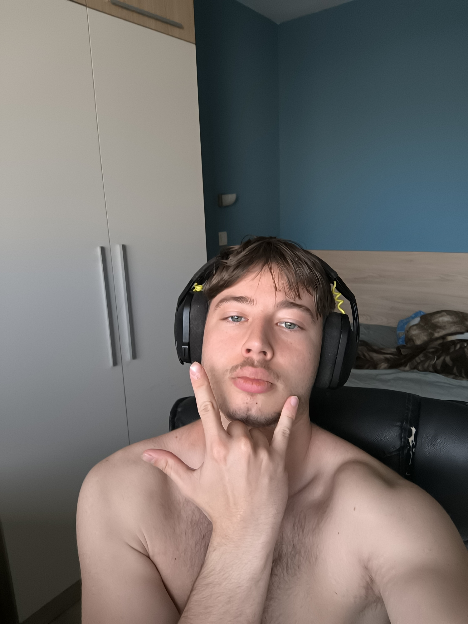 A shirtless, fair-skinned young man is seated in a black leather chair, wearing black headphones with yellow accents. He is making the "rock on" hand gesture. His eyes are blue, and he has short brown hair and light facial hair. The background includes a white wardrobe with silver handles, a blue wall, and part of a bed with blankets. The location in the city of Sofia, Bulgaria cannot be determined.