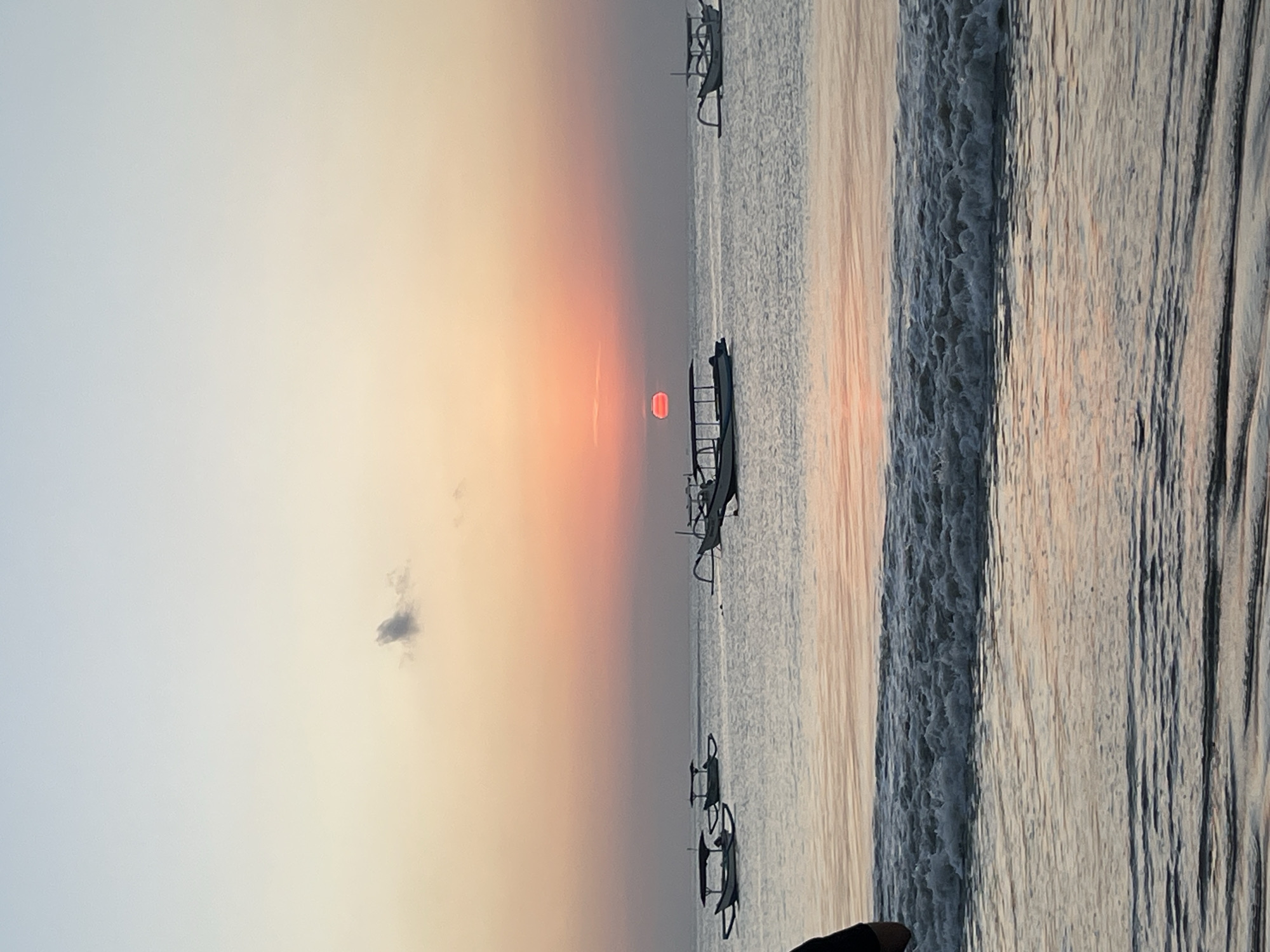 The image depicts a serene beach scene at sunset in Kuta Selatan, Indonesia. The sky transitions from a pale blue at the top to soft hues of orange and pink near the horizon, indicating the sun is setting. A single, small, dark cloud is visible in the upper left portion of the sky. The sun itself is a bright orange disc positioned just above the horizon, with its reflection slightly visible on the calm water. Several traditional outrigger boats, known as "Jukung," are anchored or floating on the sea, their silhouettes outlined against the vibrant sky. The foreground shows the gentle lapping of waves on the sandy shore. A person's dark clothing is visible on the far left edge of the frame, suggesting their presence at the beach. The overall atmosphere is peaceful and tranquil, characteristic of a sunset by the ocean.