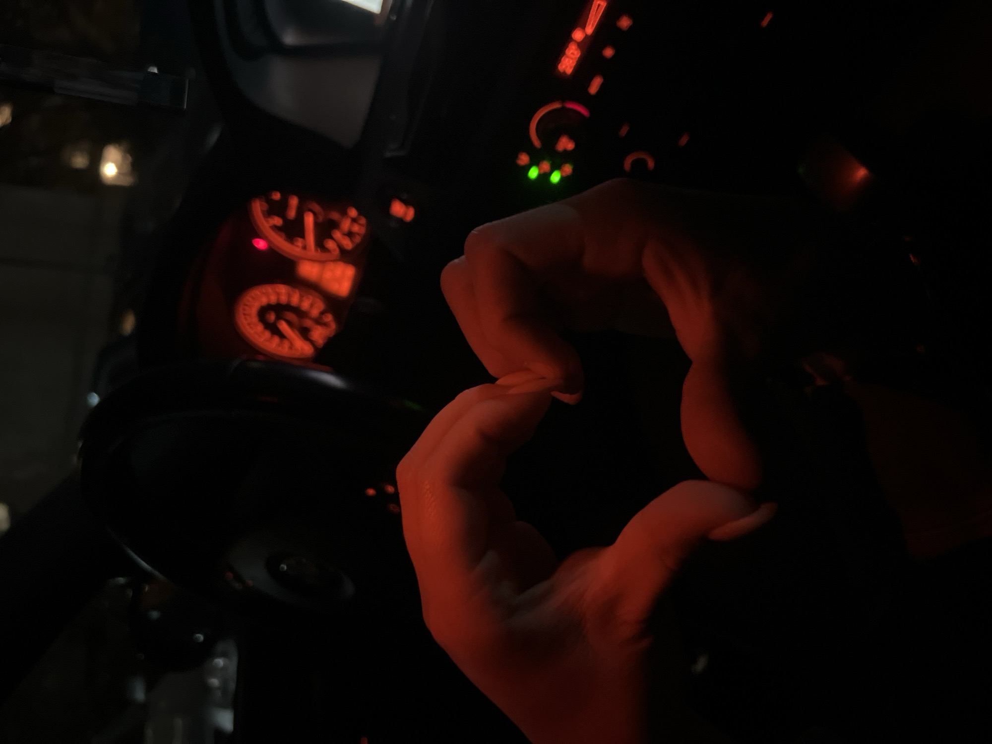 The image displays the interior of a car at night, with a primary focus on two hands forming a heart shape in the center. The hands are illuminated by a distinct red glow emanating from the car's dashboard.
The scene is set inside a vehicle at night, indicated by the pervasive darkness and the reliance on internal car lighting. On the left side of the frame, part of the car's instrument cluster is visible, featuring circular gauges with red backlighting. Numerical markings such as '20', '40', and '60' can be faintly discerned on one gauge, along with a '1/2' indicator on another, possibly a fuel gauge. A small red warning light is also active on the cluster. To the right, a section of the car's control panel shows additional red illuminated markings and three small, bright green indicator lights, likely for air conditioning or other vehicle functions.
The central activity is a person expressing affection or love by forming a heart shape with their hands. The close-up perspective of the hands suggests an intimate moment within the vehicle. The time of day is clearly night, as evidenced by the dark environment and the reliance on illuminated car controls. No specific weather conditions are discernible. The car's interior provides the sole location cue, with the broader context being Plovdiv, Bulgaria. Beyond the numerical and symbolic markings on the car's dashboard, no legible text is visible.