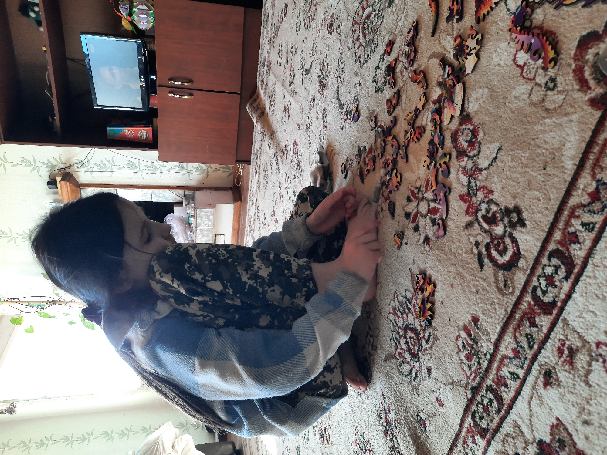 A girl sits barefoot on a patterned rug, assembling a wooden jigsaw puzzle. The rug features floral designs and a border pattern. Puzzle pieces are scattered on the rug near her. She is wearing camouflage pants and a blue and white plaid sweater. Behind her is a wall with a leafy pattern, a window with plants, and a cabinet with a television on top. The location is Архангельское, Russia.