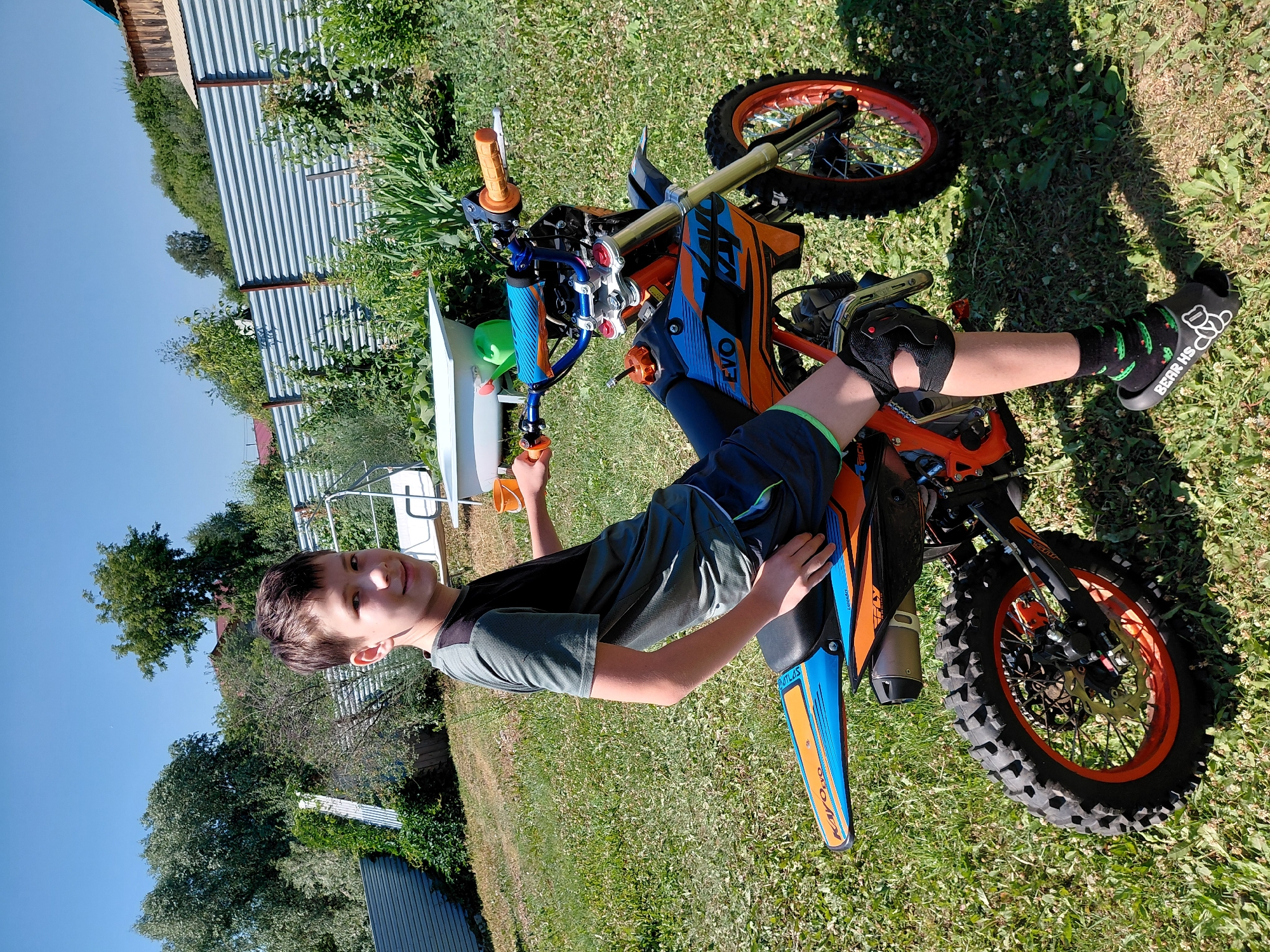 In a grassy yard, a young boy poses with a dirt bike. He is lying on his side, propped up by his arms, with his legs bent. The boy has short brown hair and is wearing a grey t-shirt and blue shorts. He has knee pads on his knees and black socks with a cartoon alligator design and the text "BEAR HS" on them.
The dirt bike is orange and blue with black tires and red rims. The front wheel is visible, as is part of the handlebars and the seat. The bike has "EVO" and "KAYO" written on it in white lettering.
The background shows a sunny outdoor scene with a corrugated metal fence, green trees, and a bright blue sky. The sun is casting shadows on the grass, indicating it is likely midday or early afternoon. The location is Novosibirsk, Russia. The overall mood appears to be relaxed and playful, with the boy enjoying his time with the dirt bike.