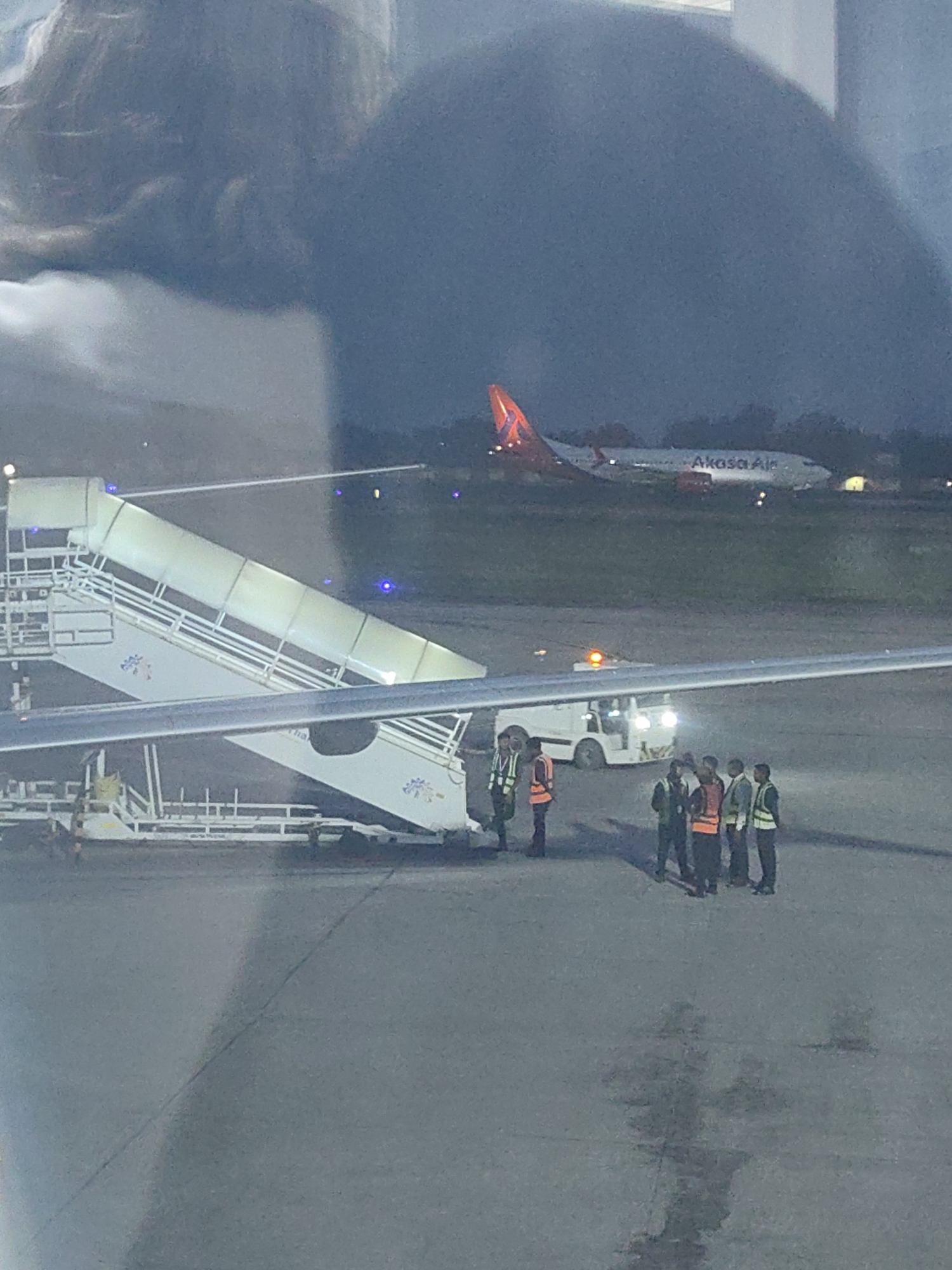 An Akasa Air airplane is parked on the tarmac at the airport in Pura Raghunathpur, India. A set of mobile airstairs is positioned next to a section of the aircraft's fuselage. Several ground crew members in high-visibility vests and one in an orange vest are standing near the airstairs and a baggage tractor. The scene is illuminated by artificial lights, suggesting it is either dusk or night.